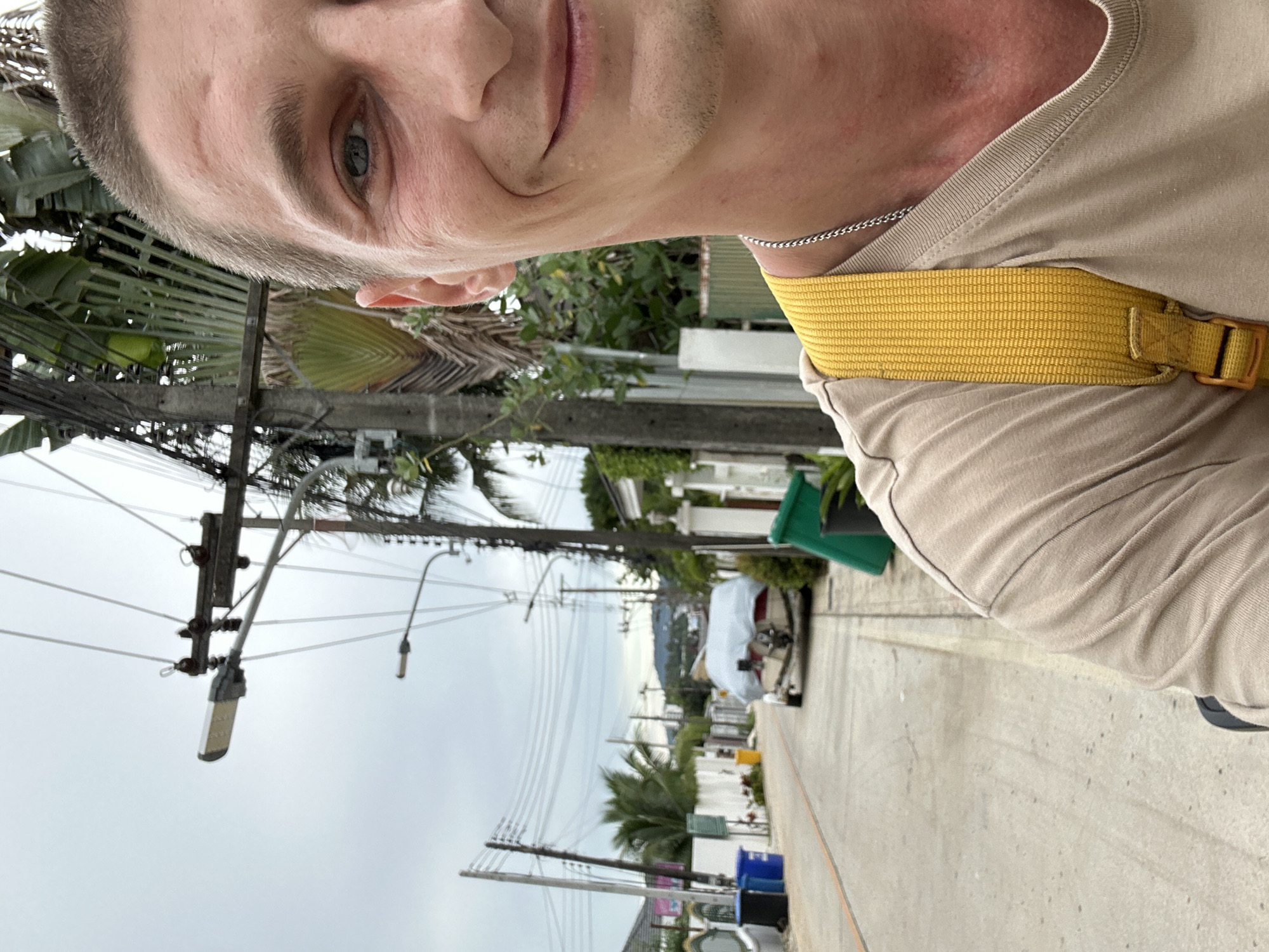 This is a selfie taken from a low angle, with the subject's face and upper body prominently featured. The person has short, light brown hair and light blue eyes. They are wearing a light beige t-shirt and a bright yellow strap across their chest, possibly part of a bag or harness. The background is blurred, but appears to be an outdoor setting with vegetation, electrical wires, and streetlights, suggesting an urban or semi-urban environment. The overall lighting indicates it is daytime, with an overcast or cloudy sky. The person's expression is neutral, and the overall mood is casual.
The image seems to be taken in Rawai, Thailand, as indicated by the presence of tropical plants like palm trees and the style of electrical infrastructure common in Southeast Asia. The ground appears to be a paved surface, possibly a sidewalk or road. In the background, there are some buildings and what looks like a covered vehicle, hinting at a residential or commercial area. The electrical poles and wires are a dominant feature, suggesting a busy street. The weather appears to be cloudy, with no direct sunlight visible. The image is oriented vertically, as if the camera was held in portrait mode. There is no visible text in the image.