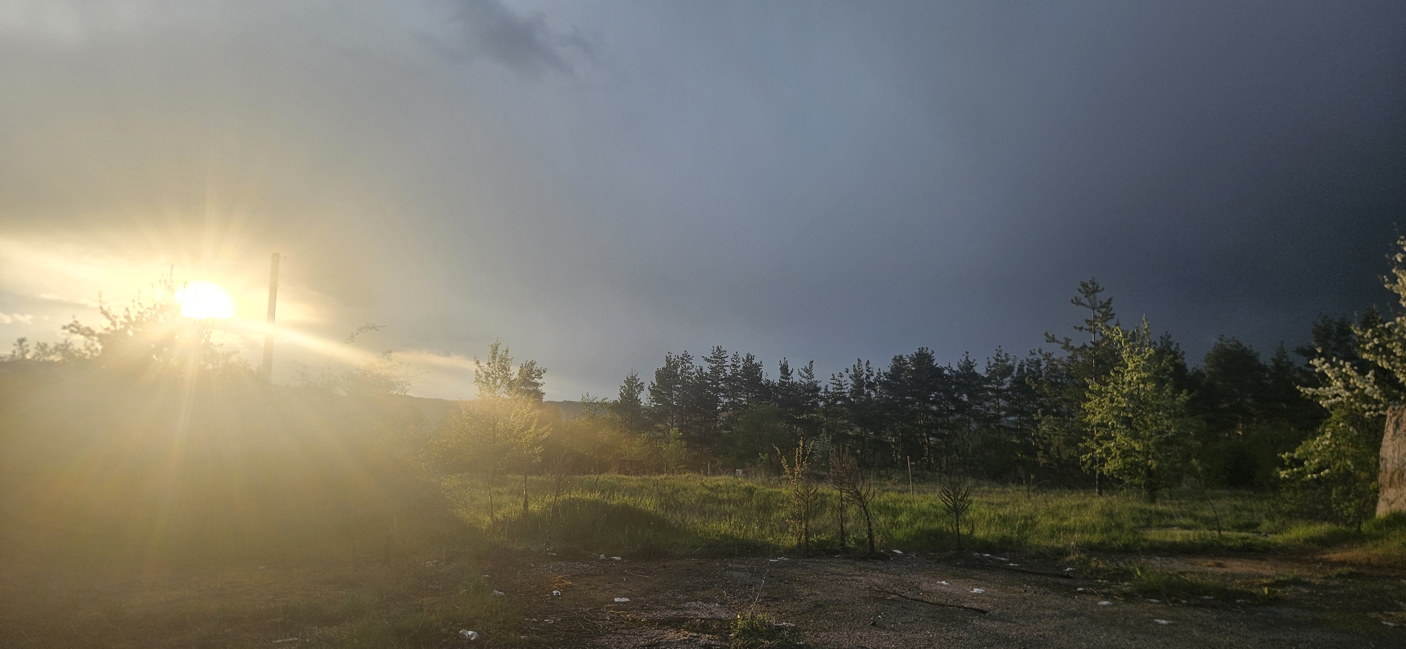 The image presents an outdoor scene with a prominent contrast between a bright, sunlit area and a dark, overcast portion of the sky. The sun is positioned on the left side of the frame, emitting strong rays and causing significant glare. A vertical pole-like object is silhouetted against the sun. In the middle ground, there's a mix of trees and vegetation transitioning from lighter greens in the sunlit areas to darker shades under the overcast sky. The sky progresses from lighter gray on the left to a dark, stormy gray on the right. The foreground appears to be a dirt or gravel ground with some scattered debris or small objects. The scene is likely located in Gutsal, Bulgaria based on the image request.