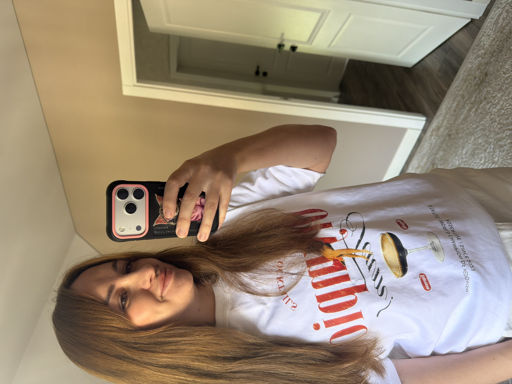 In Malinovka, Russia, a young woman with long, auburn hair is taking a selfie in a room. She is wearing a white t-shirt with a graphic design featuring red text and an image of a glass of dark liquid. Her phone, in a black case with a pink accent and a cat design, is visible in her hand as she captures the reflection in a mirror. The background reveals a doorway with two doors, one ajar, leading to another room with a wooden floor. The walls are a neutral beige, and a patterned rug covers part of the floor. The lighting is soft, suggesting an indoor setting.