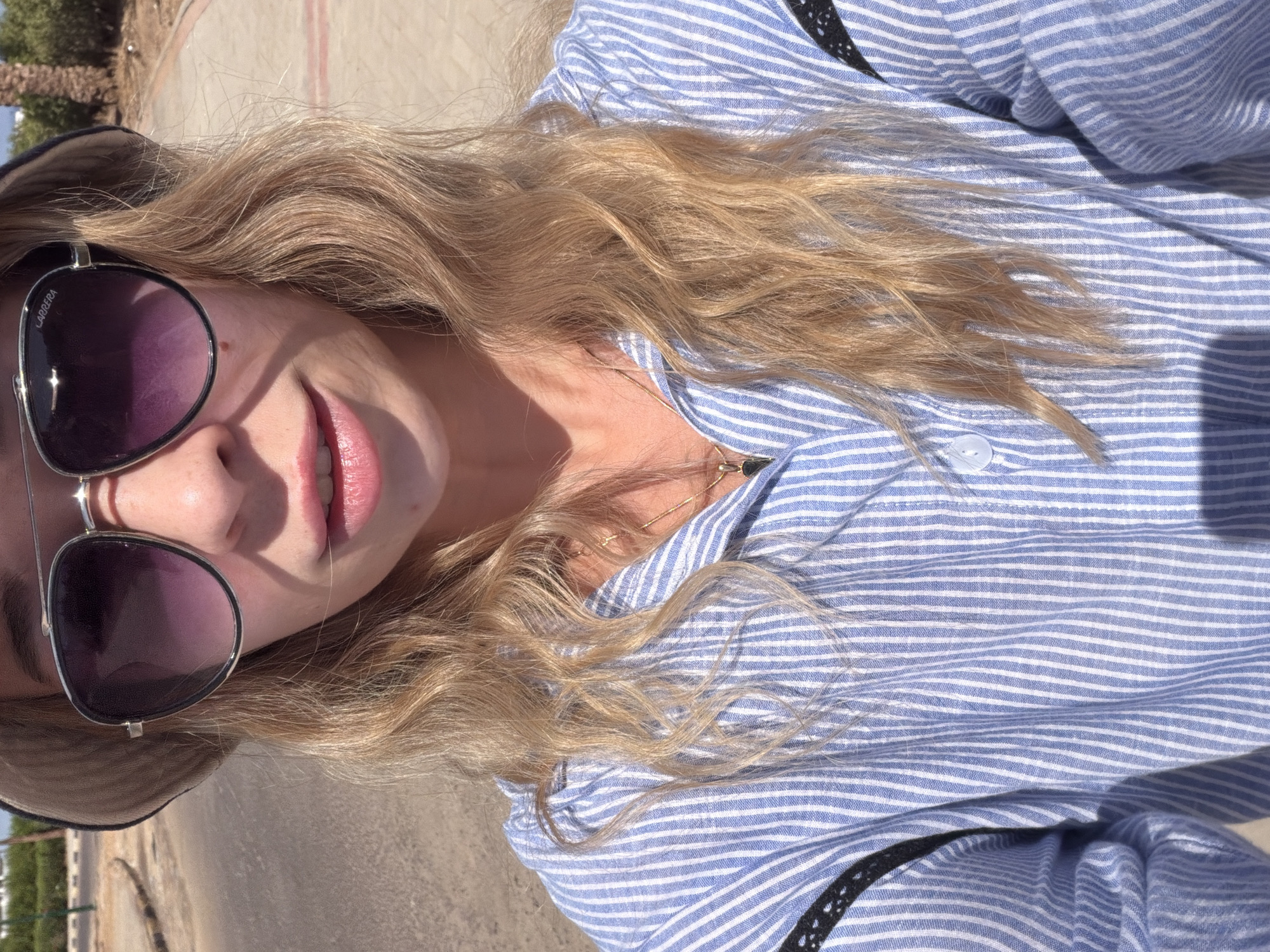 A woman is captured in a selfie, her head and shoulders filling the frame. She is wearing a light-colored bucket hat and large aviator sunglasses with purple lenses. Her blonde hair is wavy and falls around her shoulders. She is wearing a blue and white striped button-down shirt. The background appears to be an outdoor, urban setting, possibly a street in Sharm El Sheikh, Egypt, with a paved walkway and a dirt area visible. The lighting suggests it is daytime, with some shadows indicating sunlight.