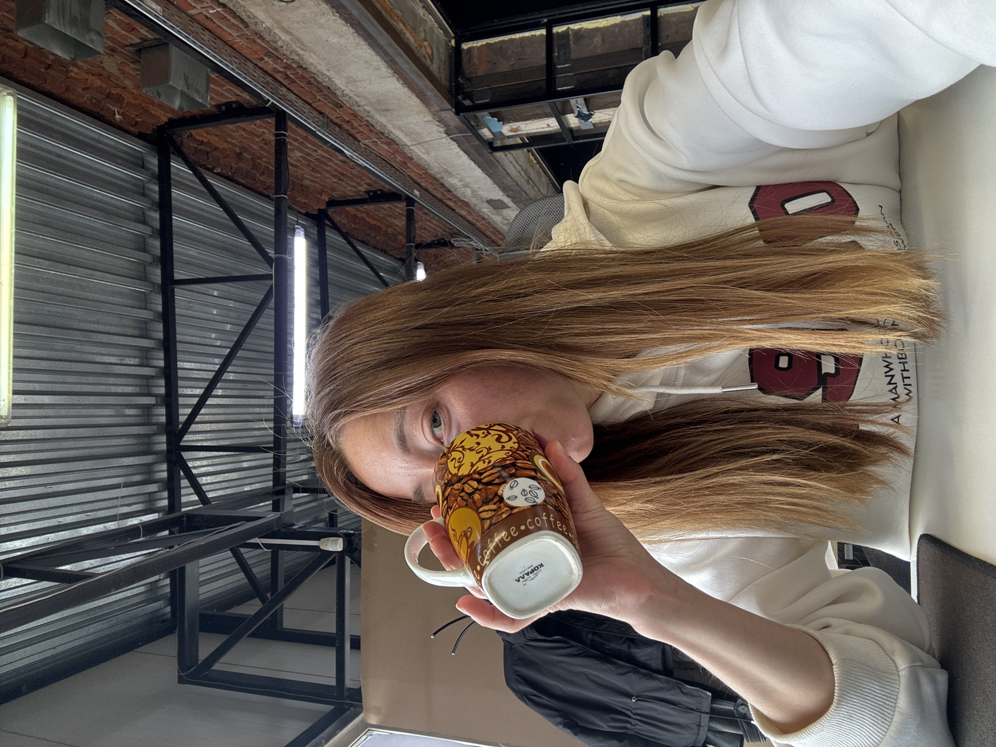 A fair-skinned young woman with long brown hair is drinking from a mug with the word "coffee" and coffee beans on it. She is wearing a white hoodie with an academic logo. The location is an industrial-style building in Chaikovsky, Russia. The walls are made of corrugated metal and the ceiling has exposed brick.