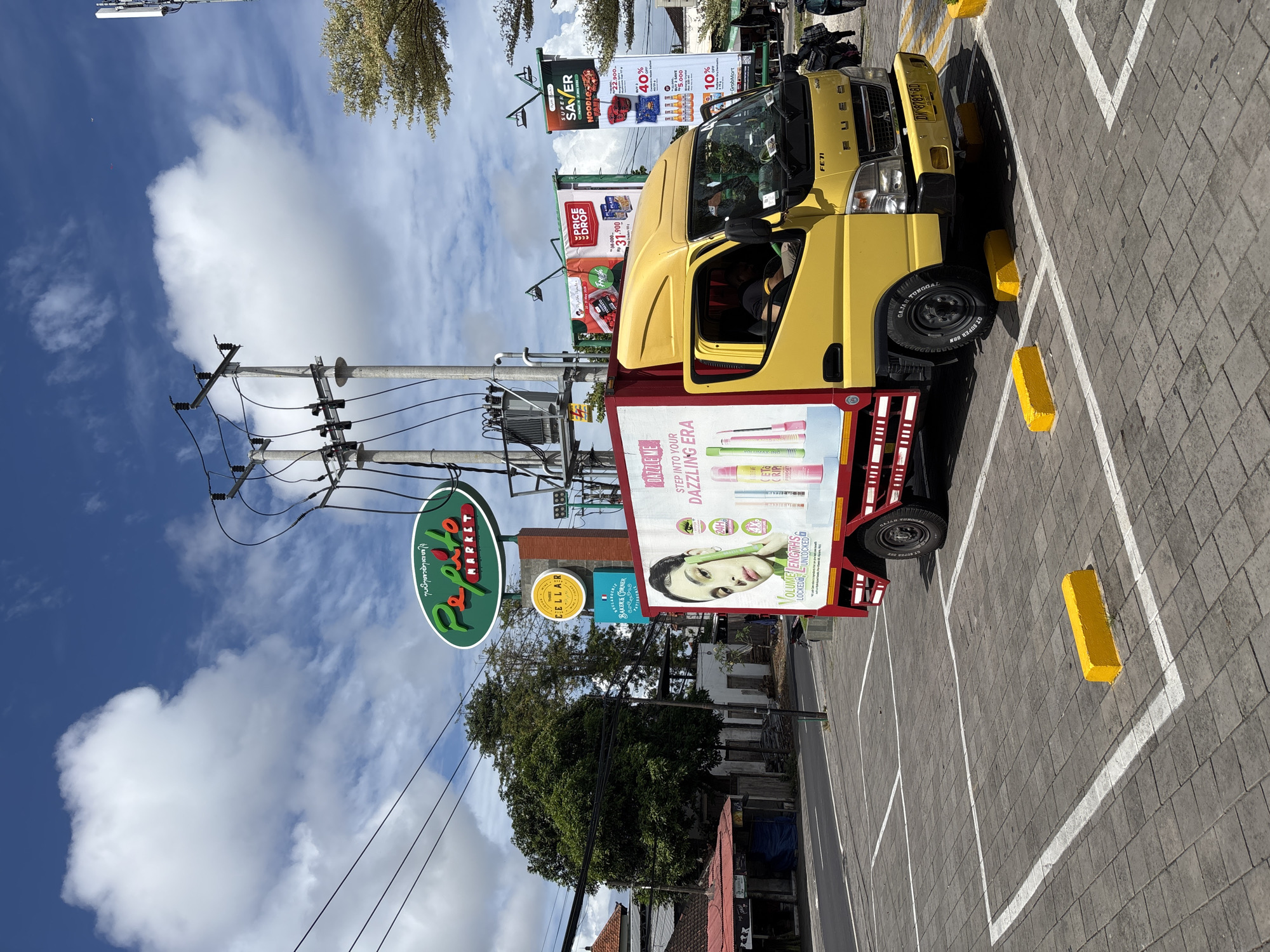 The image shows a yellow truck parked in a parking lot in Kuta Selatan, Indonesia. The truck has an advertisement for "DAZZLE ME" cosmetics on its side. A "Papito Market" sign is visible above the truck. Additional signs for "THREE CELLAR," "BOULANGERIE BAKERS CORNER PATISSERIE," and GrobMart are also visible. The parking lot is paved with light grey stone bricks and marked with white lines. Yellow concrete parking blocks are also present. Banners advertising "SUPER SAVER NOODLES" and "PRICE DROP" are also visible. Power lines and utility poles are overhead. Trees and a blue sky with clouds are in the background.
