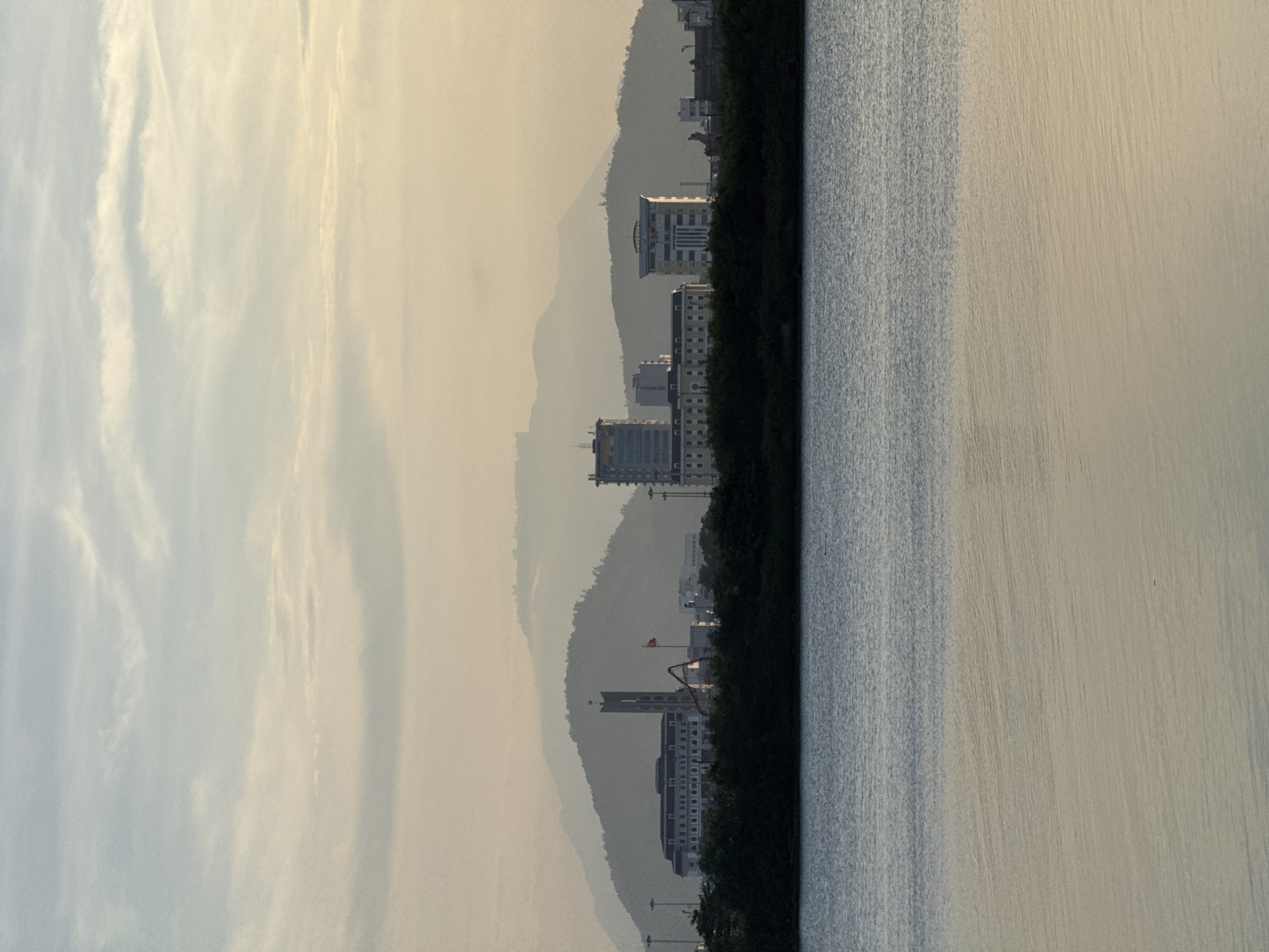 This is a wide, horizontal, landscape-oriented photograph of the Da Nang bay. The image is dominated by a calm body of water in the foreground, reflecting the soft, hazy light of either sunrise or sunset. In the middle ground, a tree line runs horizontally across the frame, separating the water from a line of buildings and hills. The buildings appear to be modern, multi-story structures, with one prominent building featuring a tall, slender tower topped with a star. Behind the buildings, several layers of rolling hills fade into the distance, silhouetted against a hazy sky. The sky itself is a pale, washed-out blue with wispy clouds, suggesting atmospheric haze or the effects of the low sun. There are no people or discernible actions in the image. The overall impression is one of a quiet, somewhat muted urban landscape viewed from across the water.