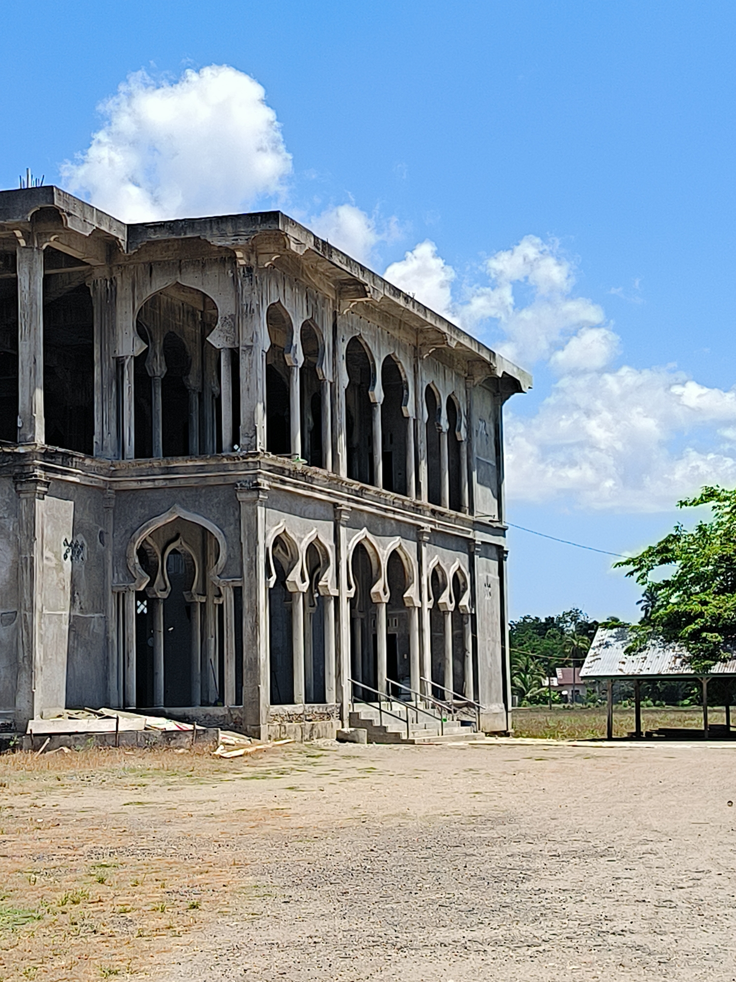 The image depicts an unfinished two-story building with architectural features reminiscent of Moorish or Islamic design. The structure is characterized by a series of arches supported by slender columns on both floors. The building appears to be constructed of concrete and is in a state of disrepair or incomplete construction, with visible signs of weathering and unfinished surfaces.
In front of the building, there is a large dirt or gravel-covered area with sparse vegetation. To the right of the building, a simple structure with a corrugated metal roof can be seen, partially obscured by trees. Beyond this structure, there is a glimpse of a residential area with houses and trees. The sky is blue with scattered clouds.
Based on the prompt information, this scene is located in Pucokalue Barat, Indonesia. However, there are no individuals visible in the image, nor is there any notable activity or interaction taking place within the frame.