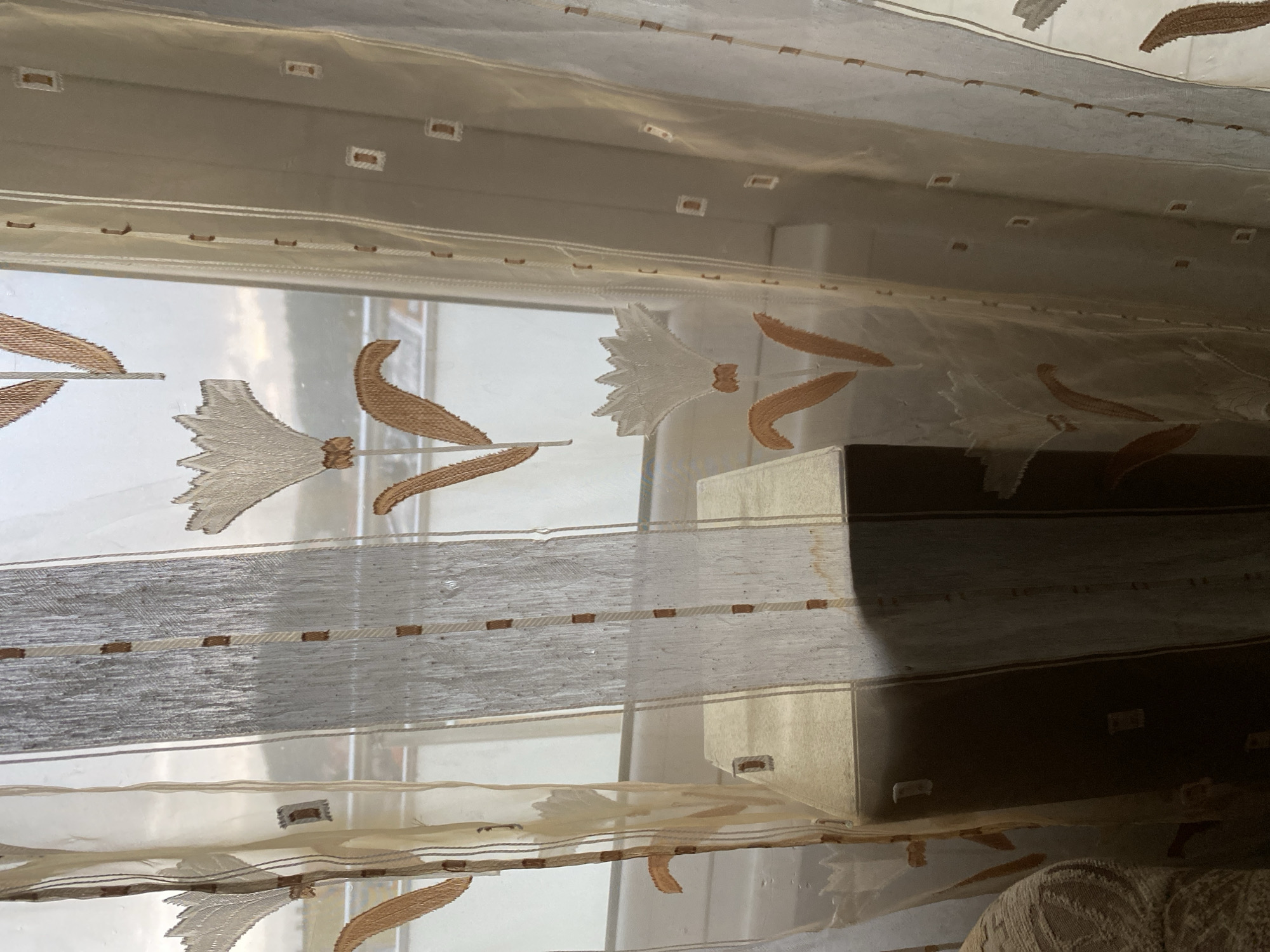 This image shows a sheer curtain with floral patterns and small squares. A brown speaker is partially visible behind the curtain. The scene appears to be indoors, near a window, with an overcast sky visible outside. There is no indication of the specific location within Bílina, Czechia.