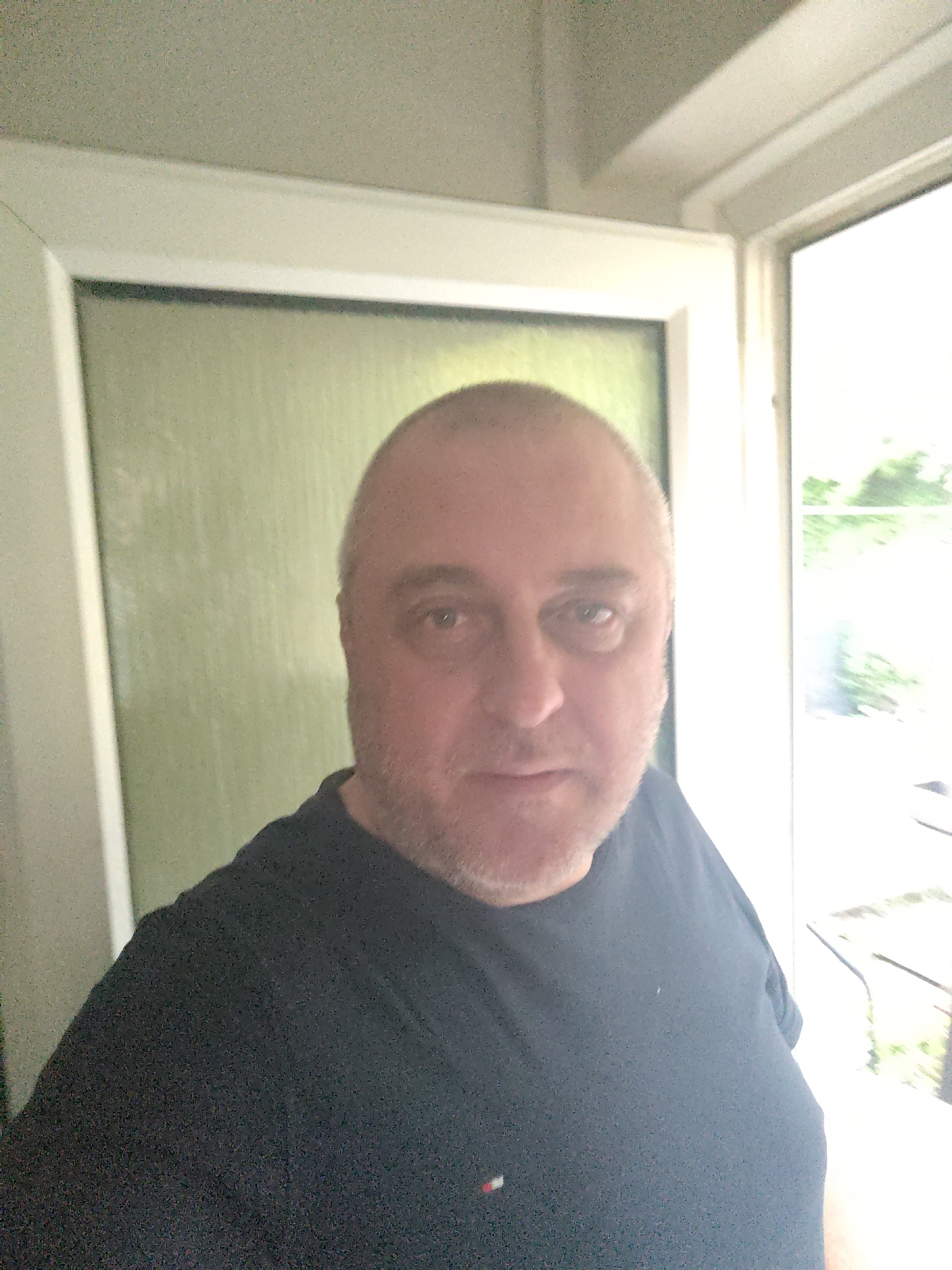 A male individual, appearing middle-aged, with a balding scalp, short light hair on the sides, and light stubble on his face, looks directly into the camera. He is wearing a dark, possibly black or navy, crew-neck t-shirt featuring a small rectangular logo with white, red, and blue stripes on the left chest area. The man is positioned indoors within the city of Huntingdonshire, United Kingdom. To his left, a white frame is visible, bordering a light green wall or frosted glass panel. To his right, a brightly lit window with a white frame shows blurred green foliage outdoors. The background elements indicate an interior space with a view to both an internal feature and an external natural environment, illuminated by daylight.