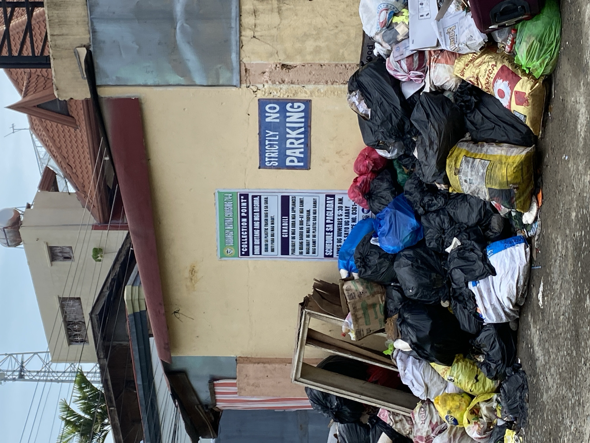 This image depicts a significant accumulation of domestic and miscellaneous refuse piled on a paved surface, obstructing a building wall in Davao City, Philippines. The scene is identified as a designated "COLLECTION POINT" for "BARANGAY MATINA CROSSING 74-A," as indicated by a green and white sign on the wall. This sign provides instructions in a local dialect, advocating for proper waste segregation, specifically mentioning placing trash in plastic bags or sacks ("ISULOD SA PLASTIC TRASH BAG O SA SAKO") and outlines a "SCHEDULE SA PAGLABAY" (collection schedule). Adjacent to this, a blue sign with white lettering clearly states "STRICTLY NO PARKING."
The garbage pile consists predominantly of numerous black plastic trash bags, varied in size, along with yellow, white, red, and blue sacks filled with waste. Visible items within the refuse include cardboard boxes, plastic bottles, a broken wooden frame, and various unidentifiable discarded materials. The waste extends from the base of the wall onto the paved ground, forming a substantial mound.
The building behind the refuse is light beige or yellow, featuring a weathered appearance. Above this immediate structure, multiple rooftops with different materials and inclines are visible, including red tiled roofs and a flat roof with a water tank. In the upper left background, part of a tall telecommunication tower is seen against an overcast sky, with power lines crisscrossing horizontally. Green palm foliage is also visible on the left side of the upper background. No individuals are present in the image.