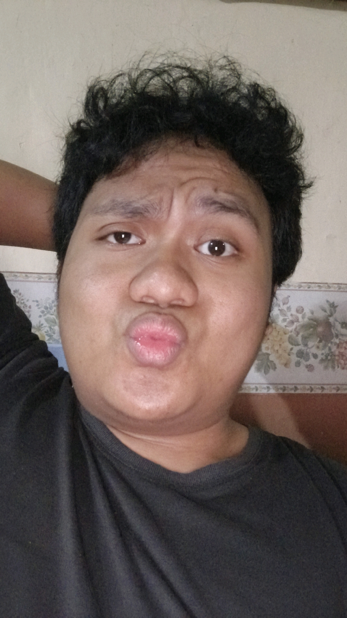 A medium shot captures a person from the chest up, posing for a selfie. The individual has dark, curly hair, and is wearing a black t-shirt. Their right arm is bent at the elbow, and their hand is placed behind their head. The person's lips are pursed in a kissing gesture, and their eyebrows are furrowed. The background consists of a wall with a floral patterned wallpaper. The lighting appears to be natural, coming from an unseen source. The image was taken in Bandung City, Indonesia.