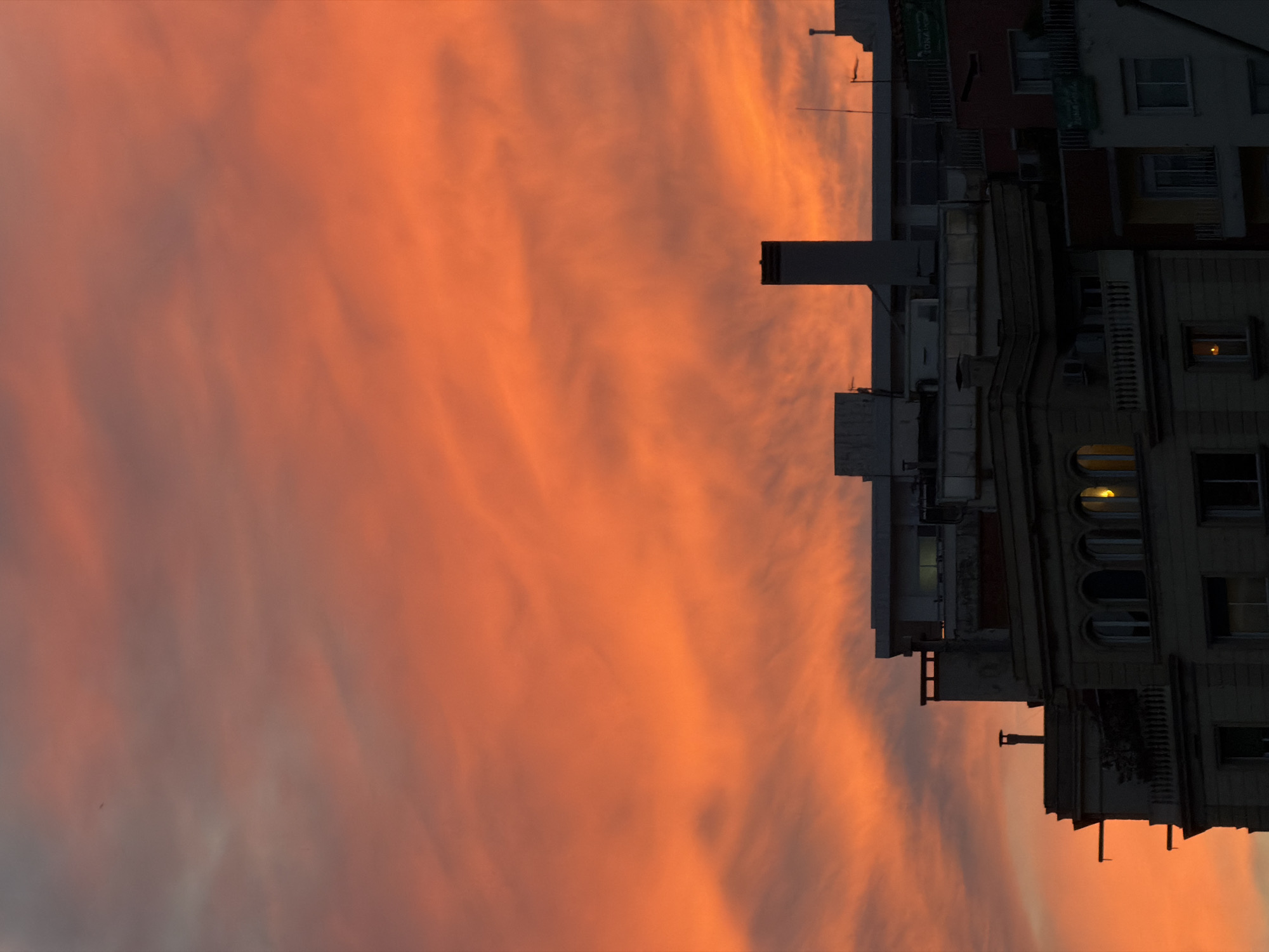 The image depicts a sunset scene in what is likely Barcelona, Spain, indicated by the architecture. The sky is filled with hues of orange and pink, suggesting either sunrise or sunset. A building's silhouette is visible on the right side of the frame, with a few windows illuminated from within. The building appears to be old and possibly residential, with a combination of dark and light elements. The image is rotated ninety degrees. No people are visible.