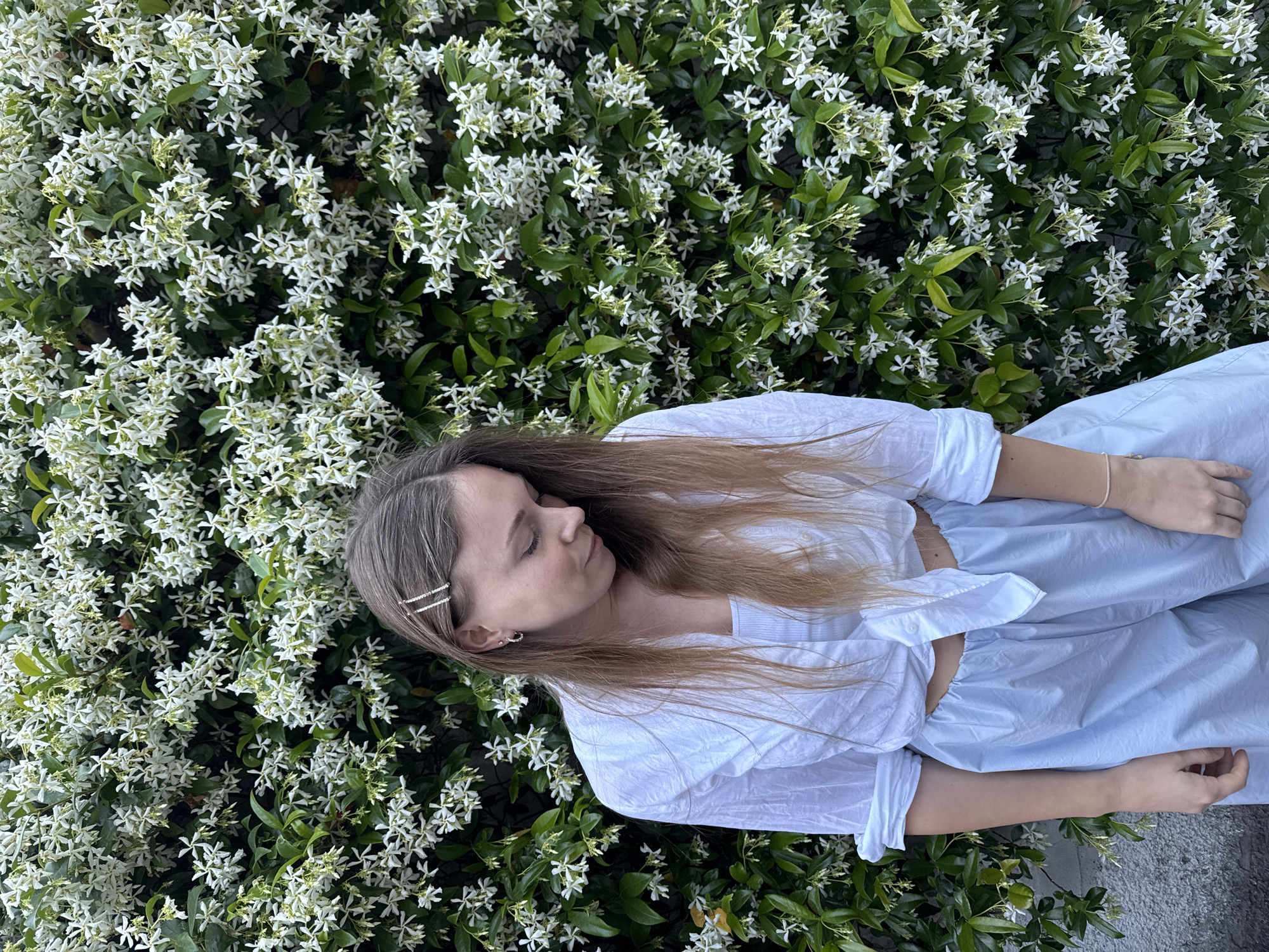 The image shows a young woman lying on a bush of white star jasmine flowers. The woman is wearing a white top and light blue pants. She is lying on her side with her eyes closed. The background is a dense bush of star jasmine flowers, with green leaves and white blossoms. The image appears to have been taken outdoors during daylight. The location cannot be determined to be in the city of Budva, Montenegro with the available information.