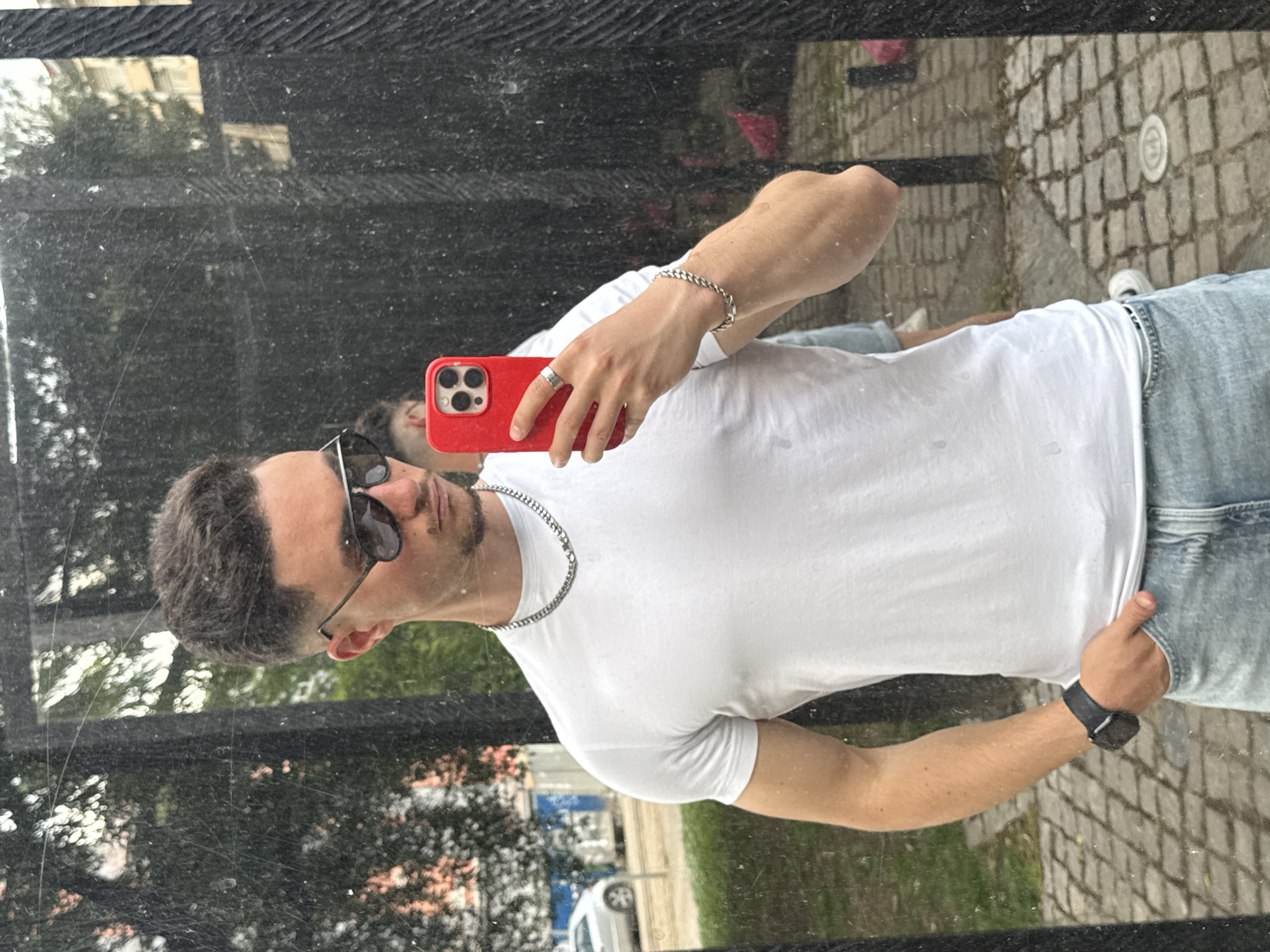 A young man with short, dark hair and sunglasses takes a mirror selfie. He is wearing a white t-shirt, light blue jeans, a silver chain necklace, a silver ring, a silver bracelet, and a dark watch on his left wrist. He is holding a red smartphone in his right hand, which is positioned to take the photo. In the reflection, another person wearing a light blue shirt is visible behind him. The man is standing outdoors, with a cobblestone ground and trees visible in the background. The environment appears to be urban, with a street and buildings in the distance, suggesting the location is in Sofia, Bulgaria.