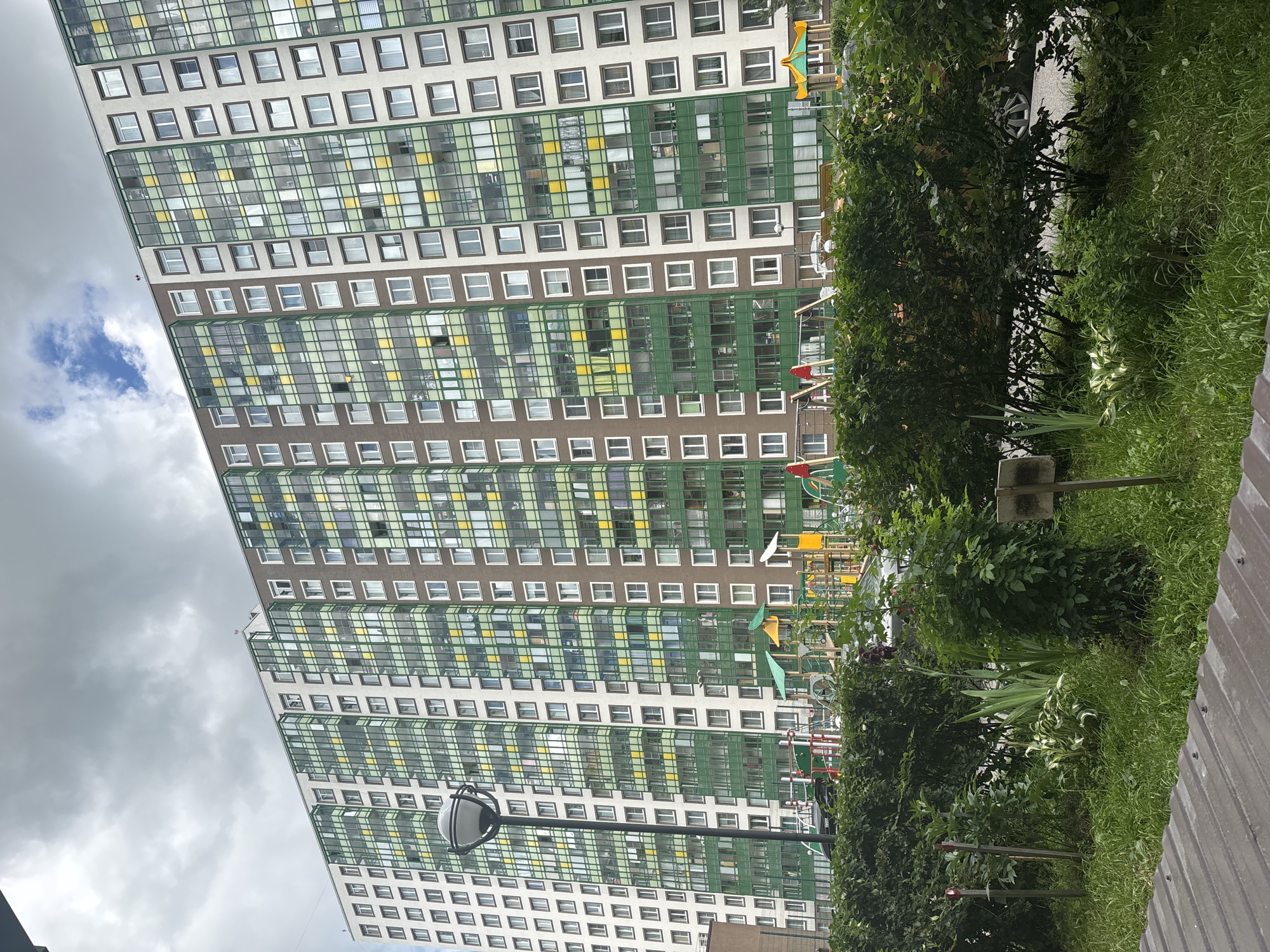 The media file shows a tall, modern apartment building with a facade of white, brown, and green sections, featuring numerous windows. The building is oriented vertically in the frame, giving a sense of its height. To the right of the building, there is a lush green area with trees, bushes, and grass, creating a contrast with the urban structure. In the foreground, a wooden or metal surface runs along the bottom right edge.
Partially visible in the background, behind the trees and along the side of the building, are elements of a children's playground, including brightly colored structures like slides and swings. A street lamp is also visible in the lower left portion of the image.
The sky is overcast with gray clouds, suggesting a cloudy day. The presence of the playground and the residential building indicates a populated area, likely a residential neighborhood. The overall scene depicts a typical urban or suburban environment with a focus on residential architecture and surrounding green spaces. There are no people clearly visible in the frame, nor any discernible text. The time of day appears to be daytime due to the ambient light.