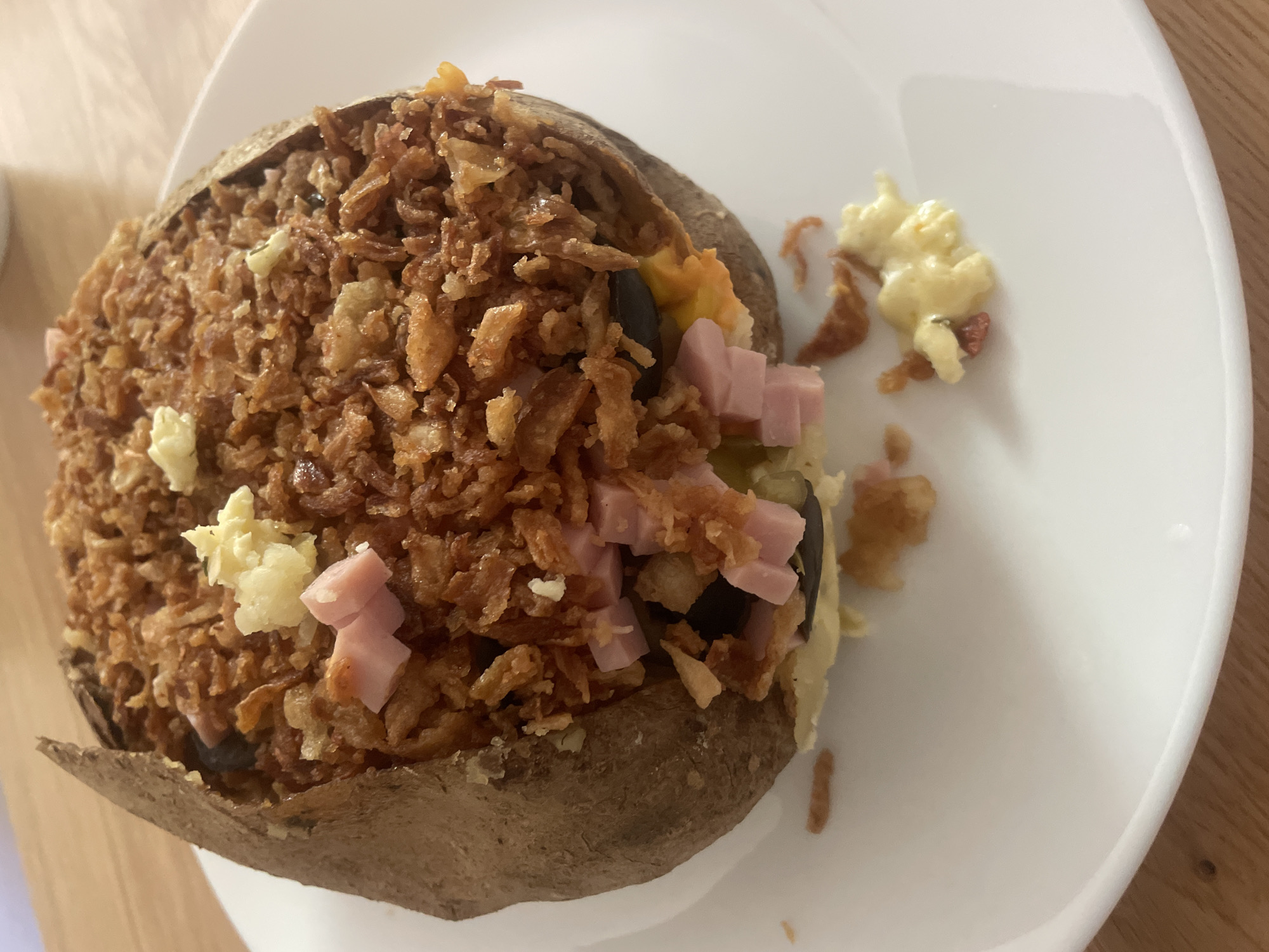 A close-up shot depicts a loaded baked potato on a white plate, situated on a wooden surface. The potato is cut open and filled with a mixture of fried onions, cubed ham, what appears to be sour cream or a similar dairy product, and other fillings. Some of the toppings have spilled out onto the plate alongside the potato. No people are visible. It is impossible to verify the location to Sofia, Bulgaria from the image provided.