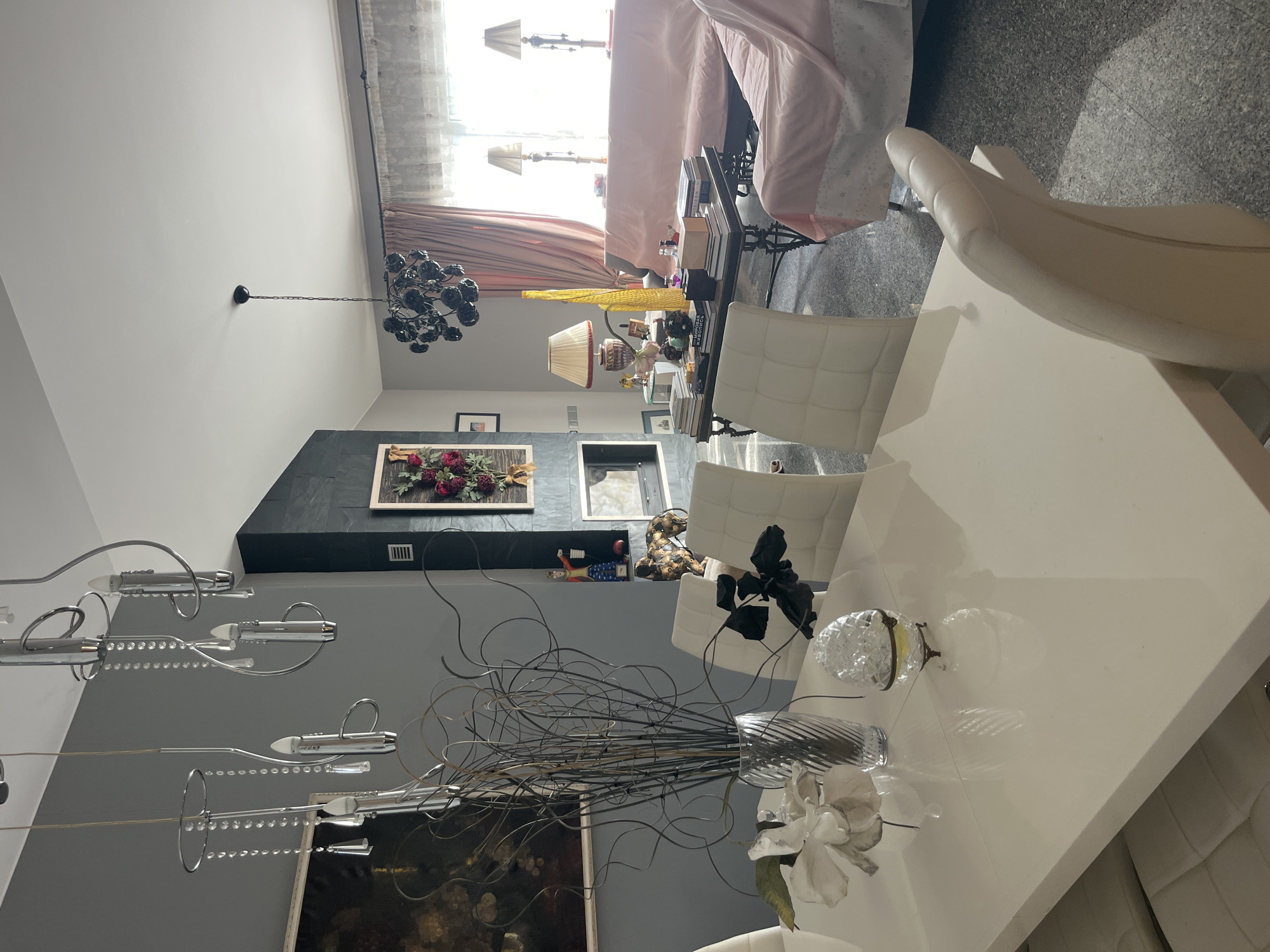 The image depicts a spacious, well-lit living and dining area in a residential setting in Parekklisia Community, Cyprus. The room features a large white dining table in the foreground, surrounded by white upholstered chairs. A decorative vase with branches and flowers sits on the table, along with a crystal egg-shaped ornament.
The living area is visible in the background, with a dark fireplace on a slate-tiled wall. A large abstract painting hangs on the gray wall to the left of the fireplace. A dark, multi-tiered chandelier hangs from the ceiling above the living room.
Furnishings in the living area include a coffee table laden with books and decorative items, a light pink sofa covered with a lighter pink throw, and another sofa section partially visible to the right, also with a pink throw. Two bedside lamps with cream-colored shades are positioned on side tables behind the sofa. Curtains with a floral pattern and a sheer overlay cover a large window.
The flooring appears to be speckled gray tile. The overall impression is one of a modern, elegantly decorated interior, with a focus on light colors and decorative elements. No people are visible in the image.