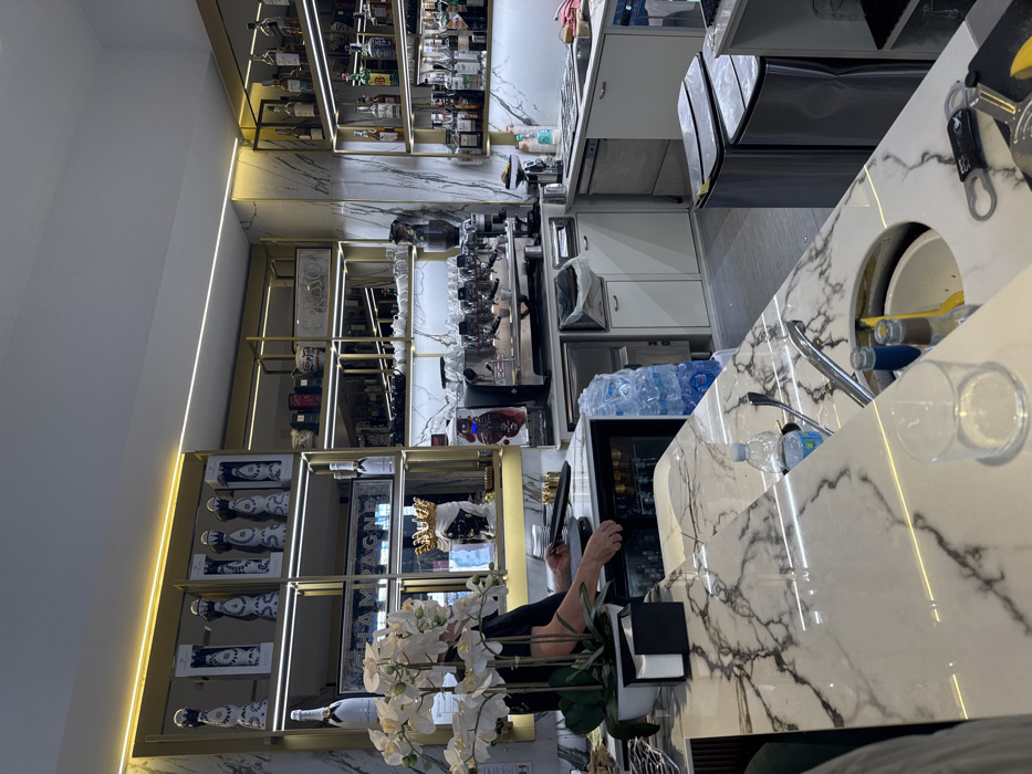 The image displays the interior of a contemporary bar or cafe, located in Milan, Italy. The environment features walls and a prominent bar counter constructed from white marble with distinct dark grey veining. Gold-toned metal shelving units with integrated linear LED lighting extend across the back wall.
The shelving on the left displays decorative blue and white ceramic bottles or vases, alongside multiple bottles of champagne, including Moët & Chandon, and a large illuminated "CHAMPAGNE" sign. A black bust wearing a gold crown is also visible on a lower shelf. Towards the right, the shelves are stocked with numerous liquor bottles, including various whiskies and liqueurs.
Behind the marble counter, a professional multi-group espresso machine is centrally positioned, flanked by smaller coffee preparation equipment, blenders, sinks, and under-counter refrigerators. A single individual, identified by their forearms and lower torso, wearing a dark shirt, is engaged behind the bar. Their left hand reaches into an under-counter refrigerator, while their right hand holds a black serving tray with stacked white saucers.
The foreground bar counter features a potted arrangement of white artificial orchids, a black napkin dispenser, an empty clear drinking glass, a plastic water bottle, and a metal bottle opener with a small lemon wedge. A circular recessed opening in the counter serves as an ice bucket or waste receptacle, containing bottles and a yellow utensil. A stack of plastic bottled water is visible next to an under-counter appliance. The floor is light grey, and two stainless steel push-top waste bins are visible on the far right.