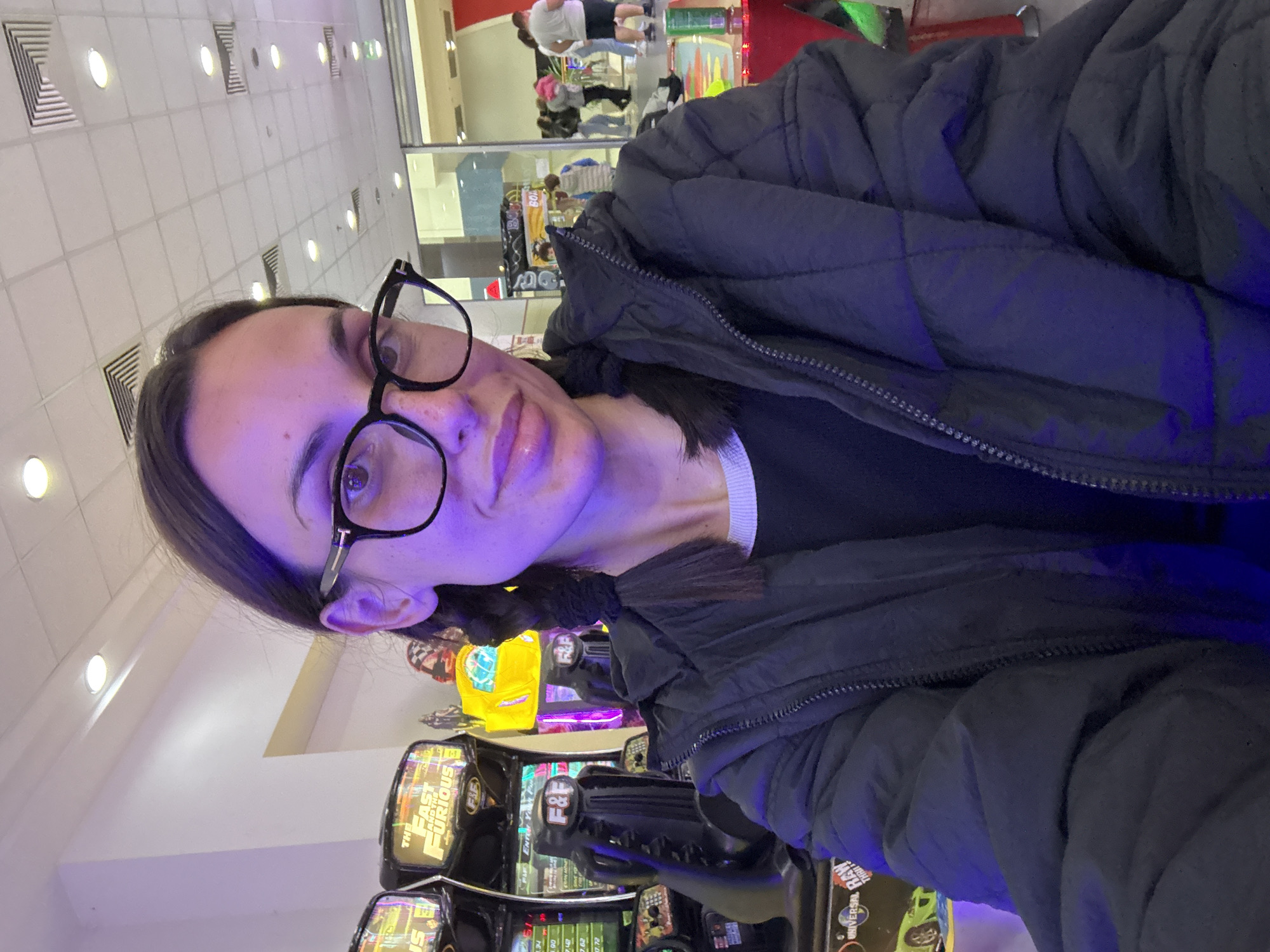 A person is taking a selfie in front of an arcade game in a brightly lit indoor location. The person, wearing glasses and a dark jacket, appears to be at an arcade game. Arcade games, including "The Fast and The Furious", are visible, including displays and steering wheels. The location is in a mall with other people in the background.