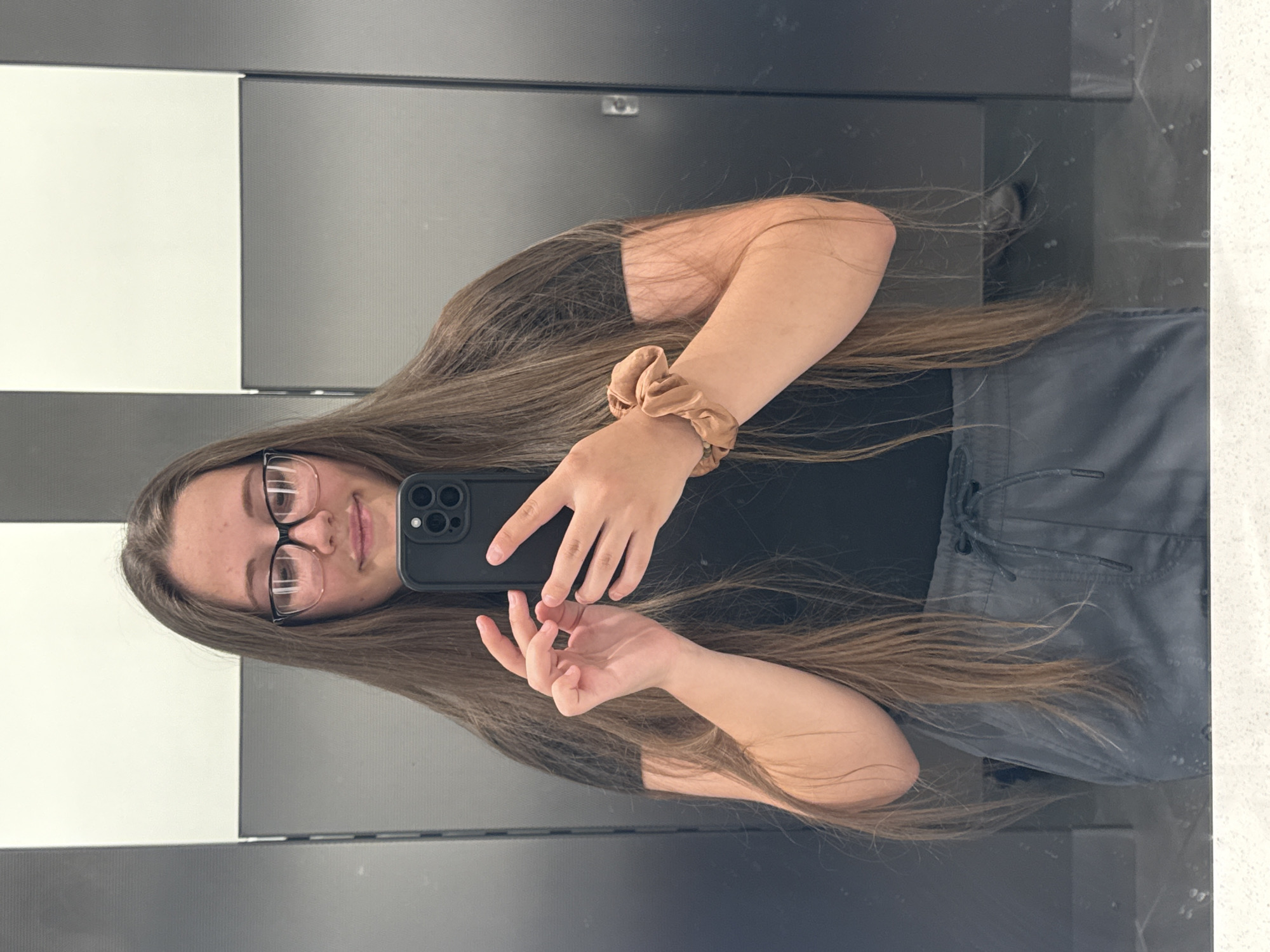 A young woman with long brown hair and glasses takes a mirror selfie in a public restroom. She is wearing a black t-shirt and grey drawstring pants. A tan scrunchie is worn on her left wrist. The background consists of grey stall doors and white partitions. There is no specific information in the image that suggests a location in Tacoma, United States of America.
