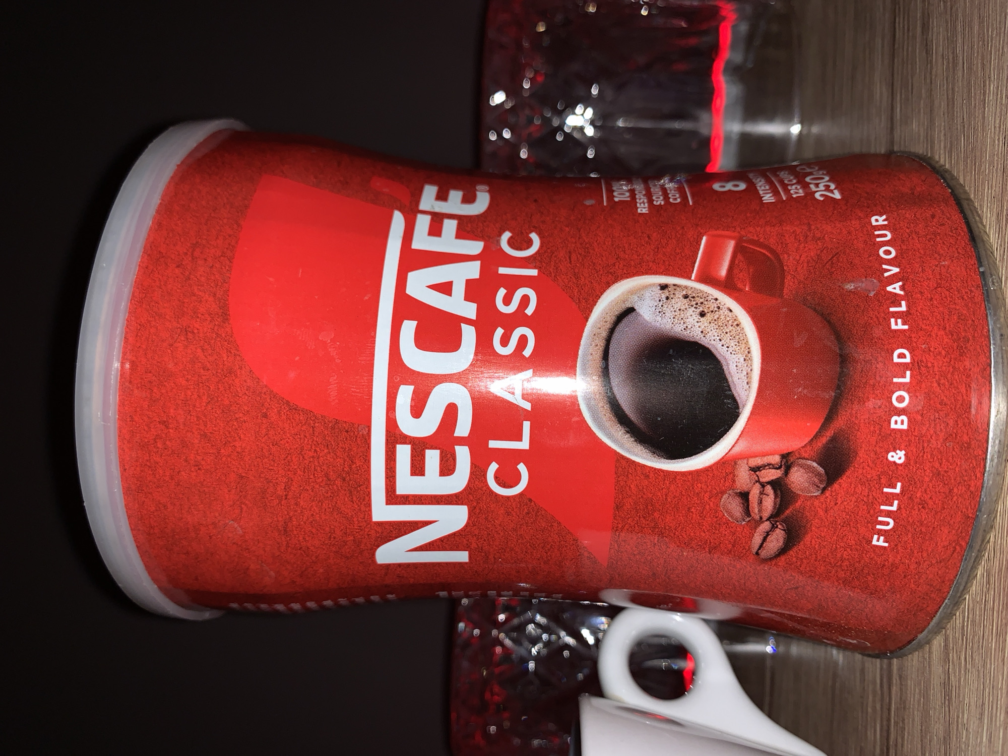 A red cylindrical container of Nescafé Classic instant coffee is prominently displayed in the foreground. The container has a white lid and a textured red surface with white lettering. The Nescafé logo is at the top, followed by "CLASSIC". Below this, an image of a red mug filled with coffee and foam is depicted, with coffee beans scattered around it. Further down, text reads "FULL & BOLD FLAVOUR". To the right of the coffee mug graphic, information such as "100% RESPONSIBLY SOURCED COFFEE", "8 INTENSITY", "125 CUPS", and "250g e" is visible. The container rests on a wooden surface. In the background, out of focus, are two glass containers with a decorative, patterned surface, reflecting light, which appear to be red in color due to the lighting. A white ceramic object with a handle, possibly a cup or mug, is visible to the left of the Nescafé container. The lighting is dim, with the primary light source highlighting the Nescafé container and creating reflections on the glass objects. The overall scene appears to be an indoor setting, possibly a kitchen counter or shelf. There is no indication of the city of Burgas, Bulgaria, in the image.