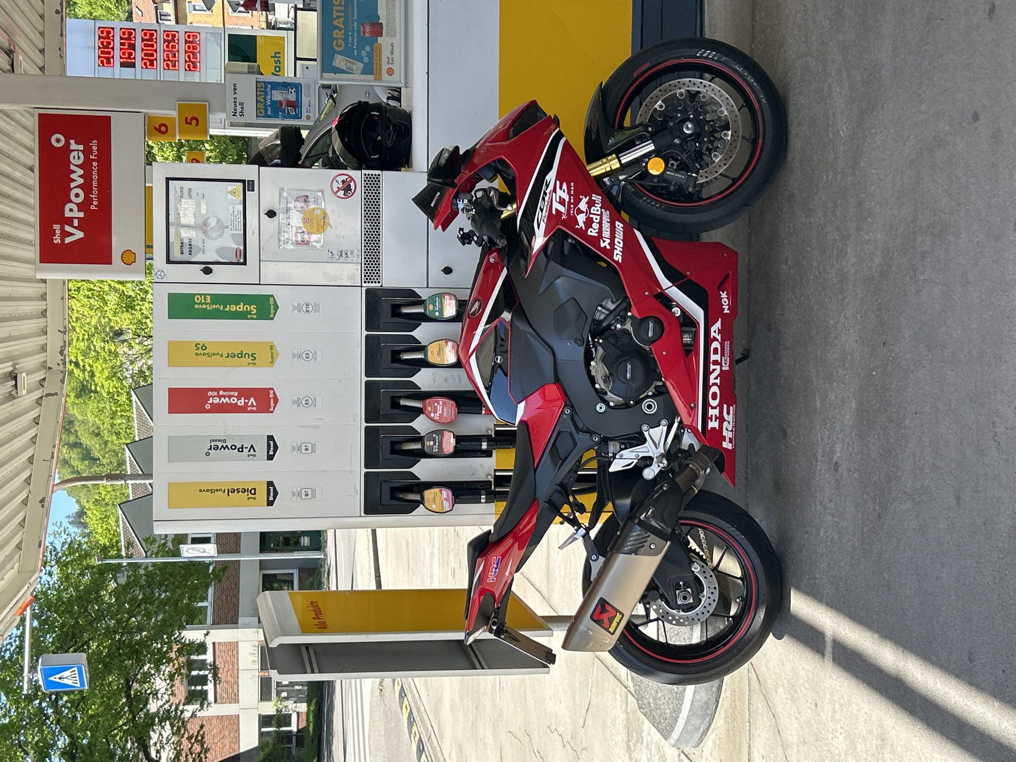 A red and black Honda CBR motorcycle is parked at a Shell gas station, seen from an overhead perspective, with the image rotated 90 degrees clockwise. The motorcycle features numerous sponsor stickers including Red Bull, Akrapovic, Showa, NGK, and Isle of Man TT, with an Akrapovic exhaust visible at the rear. A black motorcycle helmet rests on top of the gas pump unit.
The gas pump displays various Shell fuel options: V-Power Performance Fuels, Super FuelSave E10, Super FuelSave 95, Shell V-Power Diesel, and Shell V-Power Racing 100. Digital displays above the pumps show fuel prices per liter, including 203.9, 194.9, 200.9, 226.9, and 220.9 (likely Euros). Other text on the pumps and surrounding signs includes "BETRAG EURO," "ABGABE LITER," "Neues von Shell," "GRATIS zur Wäsche" (Free car wash), "GRATIS Car Wash," and "Alle Produkte," indicating a German-speaking location, specifically Eberbach, Germany.
The scene is brightly lit by the sun, casting distinct shadows on the concrete ground. In the background, green trees and buildings are visible. A blue pedestrian crossing sign can be seen in the distance. The motorcycle appears to be stopped for refueling or a brief break at the station.