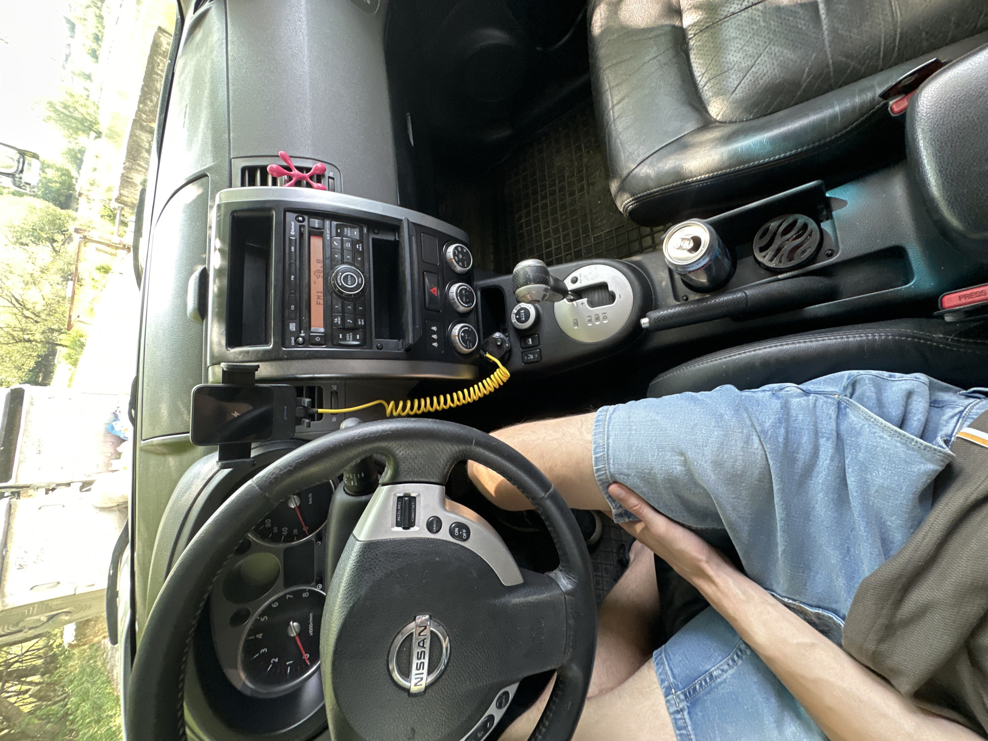 The image is a first-person interior view of a Nissan X-Trail car, taken from the driver's seat. The driver, wearing blue denim shorts, is visible from the knees down, with one arm resting on their thigh. The steering wheel, dashboard with speedometer and tachometer, radio, gear shift, and center console are prominent. A yellow coiled phone charger cable hangs from a phone mounted on the dashboard. A can of drink is placed in a cup holder on the center console. The exterior of the car is partially visible through the windshield, showing trees and what appears to be a roadside environment, possibly near Makiv, Ukraine. The lighting suggests it is daytime, with sunlight filtering into the car.