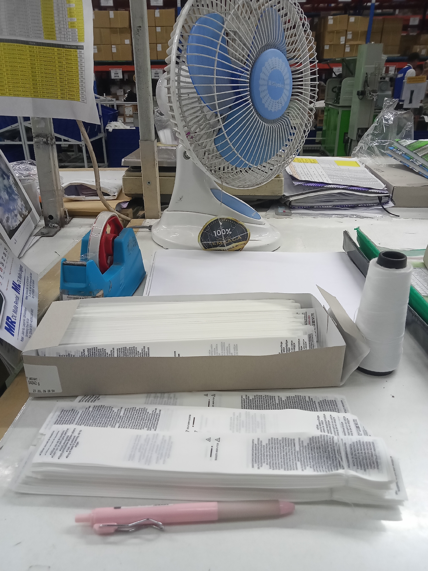 The image shows a desk with various items, including a white desk fan with a blue center, a blue tape dispenser with red tape, a cardboard box filled with white rectangular objects, a spool of white thread, and a pink pen. There are also stacks of papers and some packaging with text. The background appears to be a warehouse or factory setting with shelves and boxes. There are no people visible in the scene. The lighting is bright, suggesting an indoor environment. The overall impression is of an organized workspace, likely in a manufacturing or distribution facility.