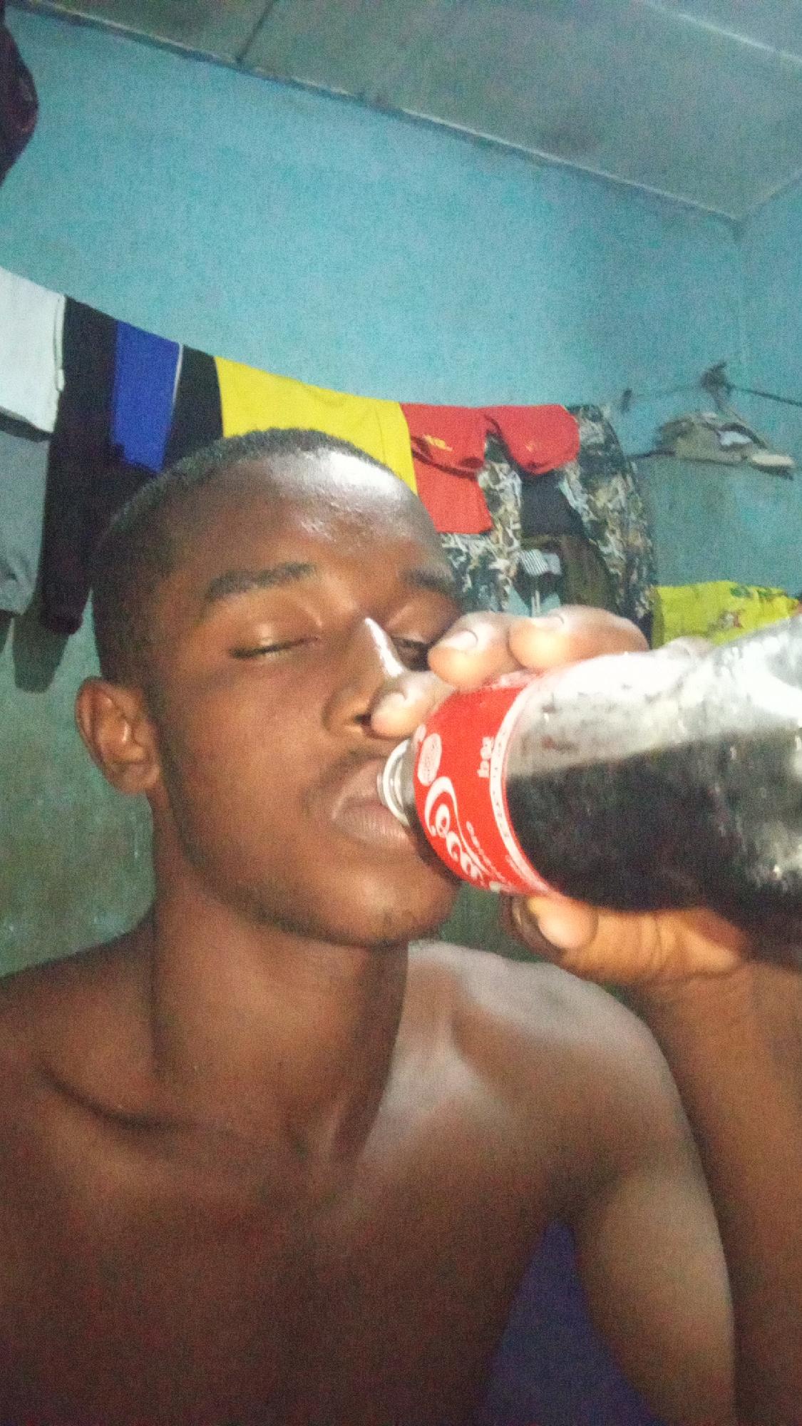 A shirtless Black man is drinking from a Coca-Cola bottle. His eyes are closed as he tilts the bottle towards his mouth. The man is indoors with a blue wall behind him. Clothes hang on a line or rod against the wall. The location is Onitsha, Nigeria.