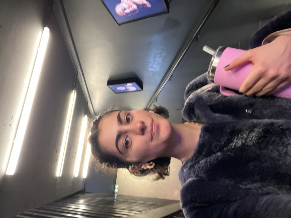 A person with dark hair holds a pink water bottle while wearing a blue fur coat. The camera angle is tilted, showing a corridor with fluorescent lights on one side and metal grates along the floor. Two framed pictures are mounted on the ceiling above the person. They have golden hoop earrings and a gold chain necklace.