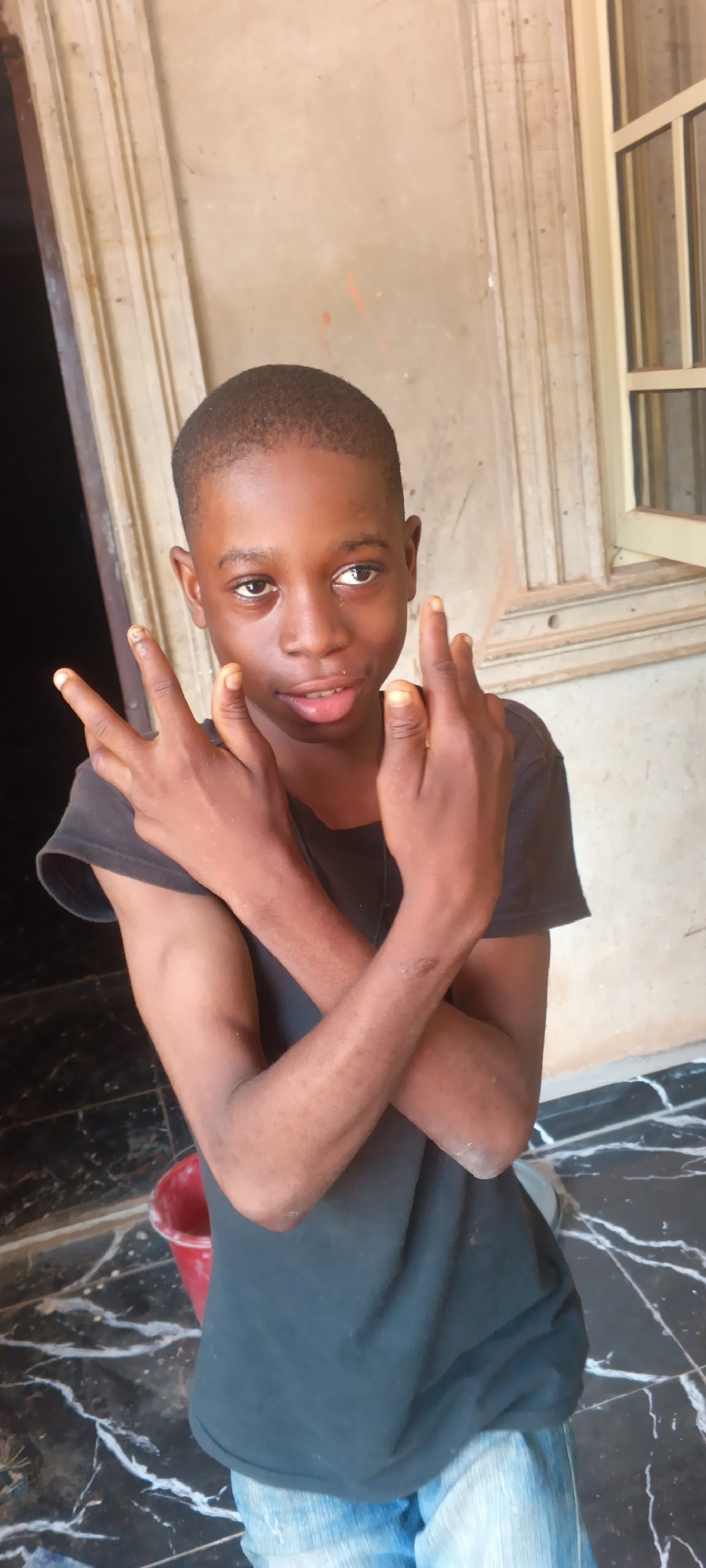 A young person stands in front of a building in what appears to be Eziudo, Nigeria. The person has dark skin, a shaved head, and is wearing a black t-shirt. The person's arms are crossed, and their hands are raised in a gesture. The building behind has a door opening on the left and a window to the right, along with marble floors. A red bucket is visible in the background to the lower left of the person.