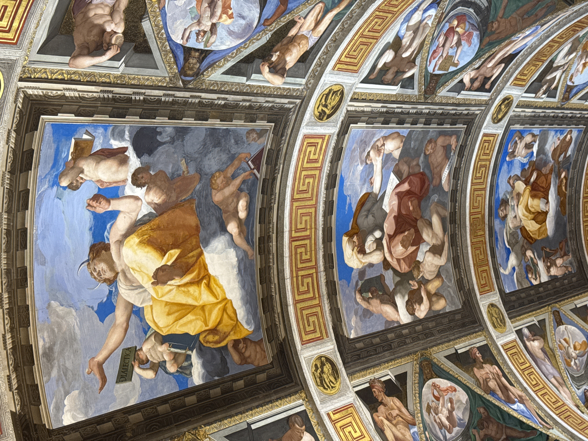 This image depicts a section of a frescoed ceiling within the Royal Site of San Lorenzo de El Escorial, Spain. The frescoes are richly detailed, featuring mythological or allegorical figures in various poses and interactions. In the upper section, a prominent female figure with a yellow garment and horns holds a tablet labeled "DIALECTICA" aloft, gesturing with her other hand. She is surrounded by cherubic figures and other human forms amidst clouds and a blue sky. Below this, a segment of the ceiling displays a Greek key pattern border above another fresco panel. This lower panel shows a group of figures, including a reclining bearded man with a yellow garment, interacting with nude and draped figures, one of whom holds a book. The overall environment is one of grandeur and artistic embellishment, characteristic of the historical and royal context of El Escorial. The artwork utilizes a vibrant color palette and masterful rendering of anatomy and drapery, conveying a sense of movement and drama.