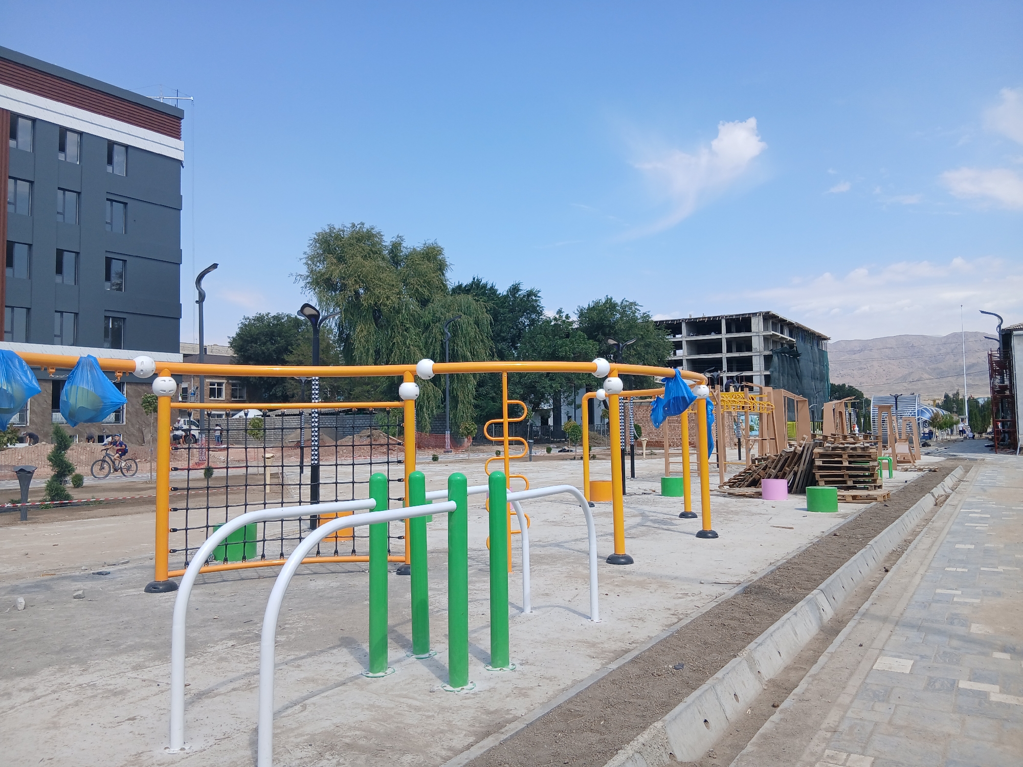 The image depicts a construction site for a public park or recreational area in Talas, Kyrgyzstan. In the foreground, there are various pieces of outdoor exercise equipment, including a pull-up bar structure with yellow uprights and white and green bars, and a black net structure. The ground is paved with concrete and dirt. To the left, a modern, grey multi-story building with a brown and white facade is visible, along with trees and streetlights. Further in the background, unfinished buildings, stacks of wooden pallets, and construction materials are present, suggesting ongoing development. A person is seen riding a bicycle on a path to the left of the exercise equipment. The sky is clear and blue with a few scattered clouds, and mountains are visible in the far distance. The overall scene suggests a developing urban area in Talas, Kyrgyzstan, with a focus on creating public amenities.