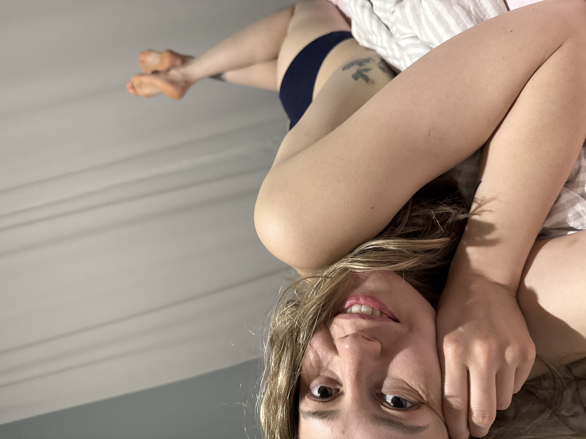 A woman is lying in bed, smiling at the camera. She has blonde hair and is wearing a dark-colored bottom. Her legs are crossed and elevated, with a tattoo visible on her thigh. The background shows a wall with horizontal lines, indicating it may be a ceiling. The location cannot be determined to be Chaikovsky, Russia, based on the image alone.