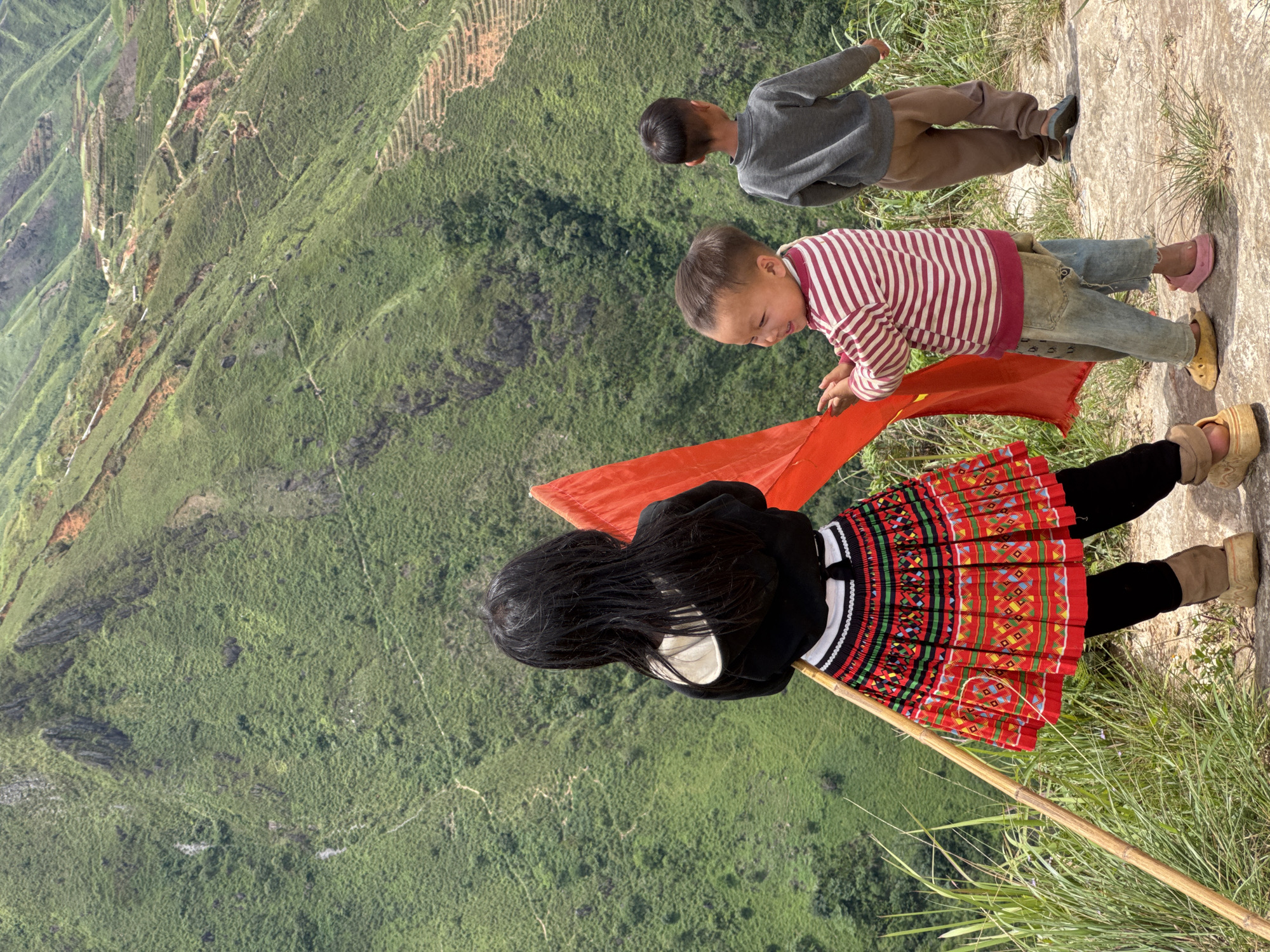 Three children are standing on a rocky, grassy ledge overlooking a vast, green mountainside with terraced fields. The child in the foreground on the left, facing away from the camera, has long black hair and is wearing a black hoodie, a colorful, pleated skirt with geometric patterns, black leggings, and large, tan, platform-like shoes. They are holding a long bamboo pole vertically. To their right, a younger child in a red and white striped long-sleeved shirt and distressed denim jeans is looking to their left, holding a large, bright red fabric. To the right of the younger child, a third child, facing away from the camera, is wearing a gray long-sleeved shirt and brown pants. The background shows a steep, lush green mountain landscape, characteristic of Ta Xua, Vietnam, with winding paths and cultivated terraces visible on the slopes. The sky is overcast, creating diffused lighting.