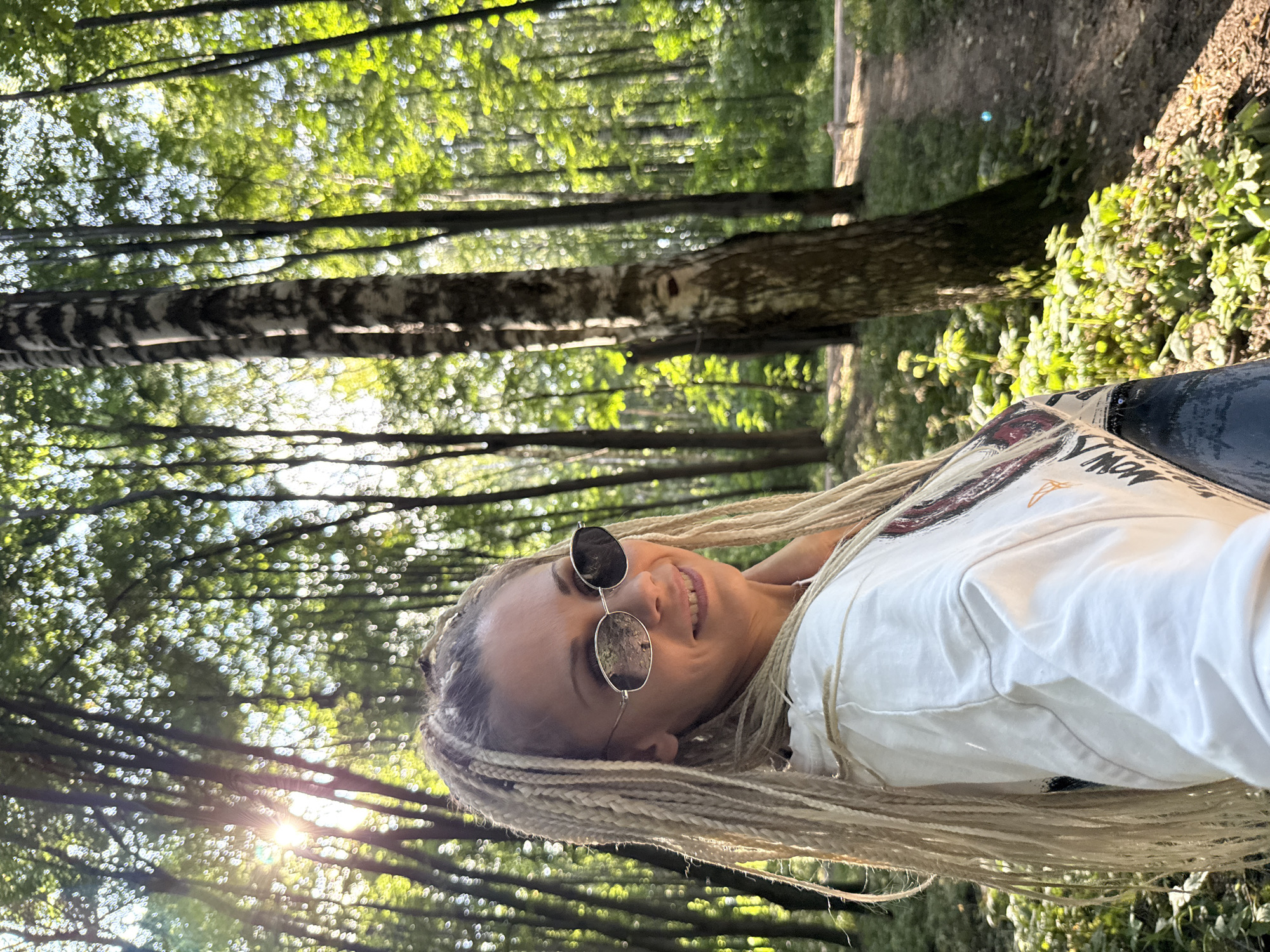 A woman with blonde braids and sunglasses smiles at the camera, lying down in a forest. She is wearing a white t-shirt with some colorful graphic design on it. The sun shines through the trees, casting dappled light on the scene. The ground is covered with leaves and some greenery. The trees are tall and spaced out, with some birch trees visible. The image appears to be taken during the daytime, likely in the late afternoon due to the warm sunlight. The woman's expression suggests happiness and relaxation. The setting is a natural outdoor environment, possibly a park or a forest trail. There is no visible text in the image.