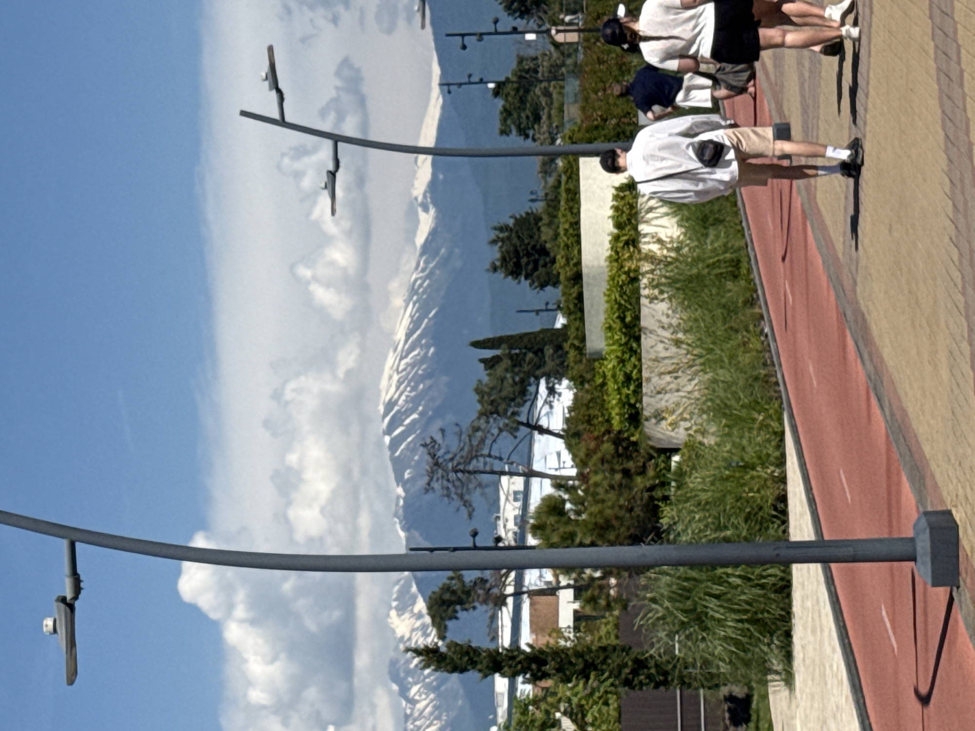 This image depicts an outdoor pedestrian and cycling area in Sirius, Russia, under a clear blue sky with scattered white cumulus clouds. In the distant background, a prominent range of mountains with significant snow cover is visible.
In the mid-ground, various types of green trees, including tall conical evergreens and more spreading deciduous trees, are interspersed with dense green shrubbery. Several modern, light-colored buildings are partially visible behind the vegetation. Further down the path, multiple dark grey streetlights with multi-arm fixtures are present.
The foreground is dominated by a dark grey, curved streetlight pole with a single, flat light fixture topped with a white sensor, positioned on the left. To its right, a distinct red asphalt track, marked with white dashed lines, runs parallel to a tan-colored paved pedestrian walkway constructed from interlocking pavers.
Four individuals are observed walking away from the camera on the paved walkway. The person closest to the left is a male wearing a white long-sleeved shirt, beige shorts, dark shoes with white socks, and a dark cap, carrying a small black crossbody bag. To his right, a male wears a dark blue short-sleeved shirt and white shorts. On the far right, a female with a dark cap and a long dark braid wears a white t-shirt, black shorts, and white shoes, carrying a dark shoulder bag. A fourth person, partially obscured behind her, is also visible in a white top and dark shorts. Shadows cast by the streetlights and people indicate bright sunlight.