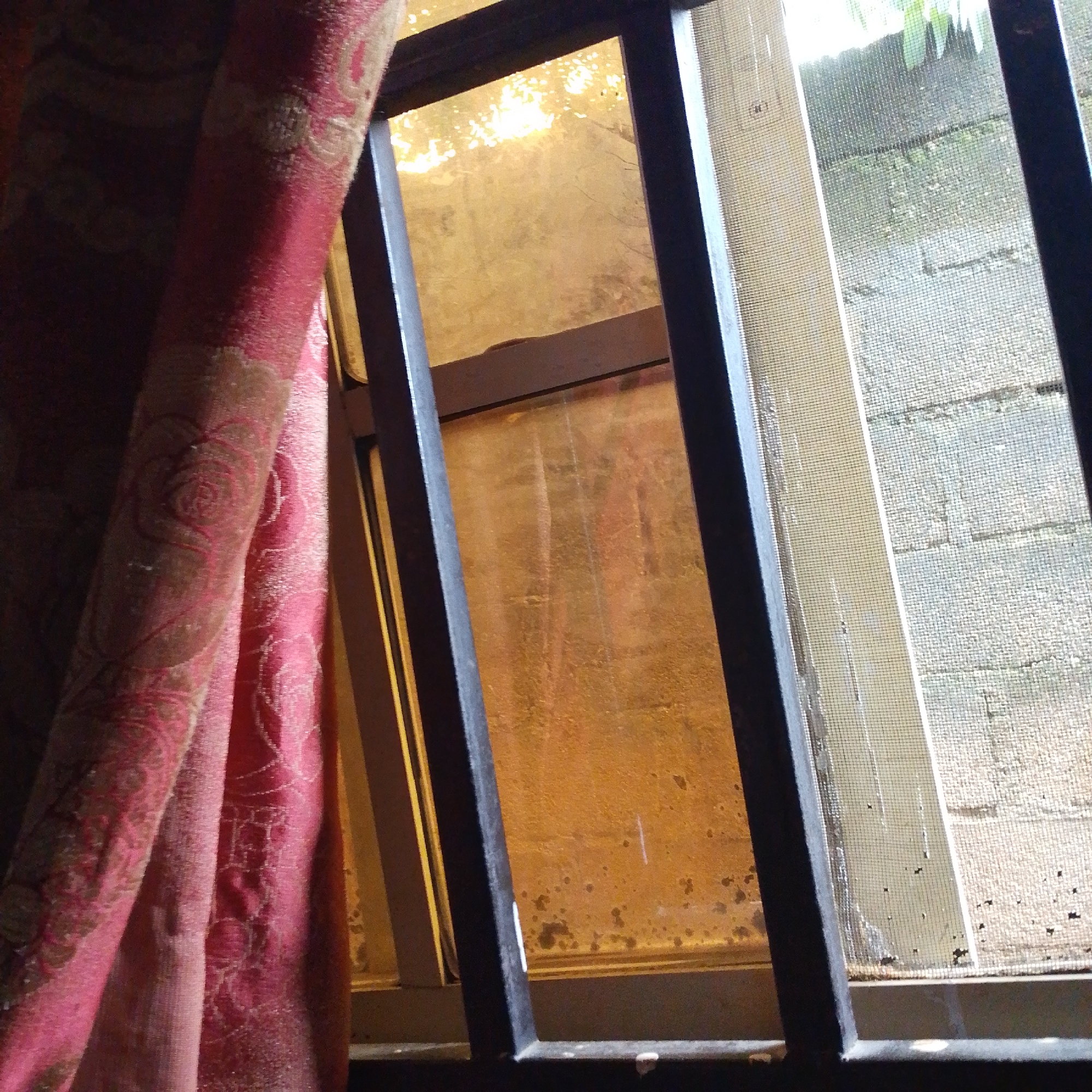 A close-up interior shot focuses on a window with a pink, patterned curtain to the left. The window is divided into two panes by dark metal framing. The left pane is covered in a translucent, yellowish material with visible dirt and water stains. The right pane is covered by a fine mesh screen, through which a textured, light-colored surface resembling a brick wall is visible. Beyond the wall, greenery can be seen. The lighting is dim, casting a warm glow from the window. The scene appears to be in Jalingo, Nigeria.