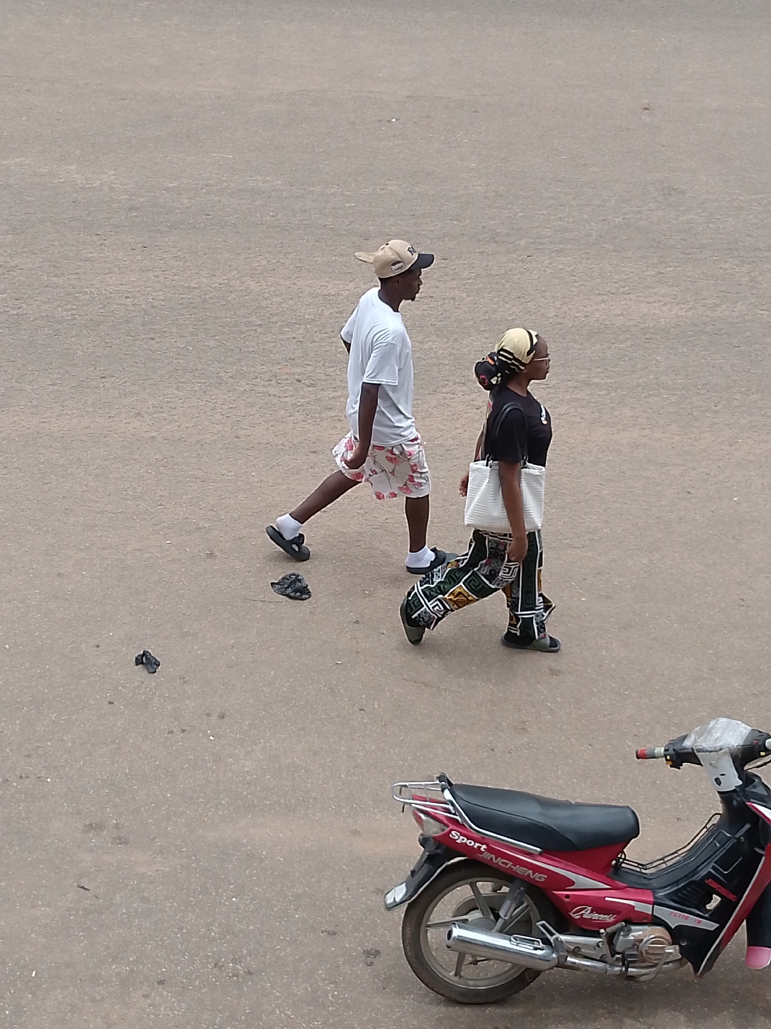 A man and a woman are walking on a paved surface in an outdoor urban environment. The man is on the left side of the image, wearing a white t-shirt, patterned shorts, a tan baseball cap, white socks, and black sandals. He is in mid-stride, with his left leg extended forward. Near his feet are two dark, crumpled objects on the ground. The woman is to the right of the man, wearing a black t-shirt, a patterned headscarf, glasses, and patterned pants. She is carrying a white tote bag on her left shoulder and is also in mid-stride, walking in the same direction as the man. In the lower right portion of the image, a red and black motorcycle is parked. The background is a flat, grey paved surface, suggesting a street or pathway. The lighting appears to be natural, possibly overcast.