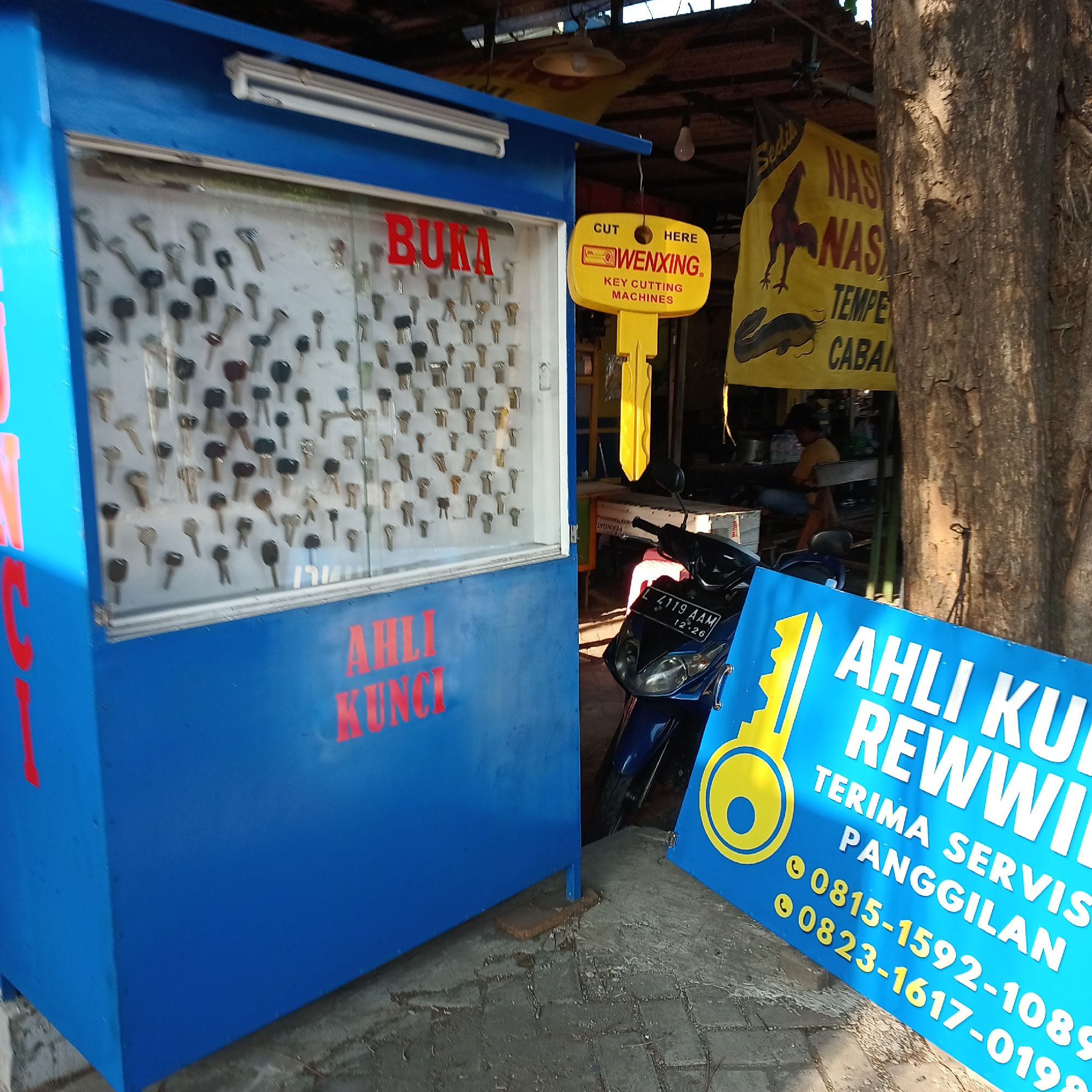 In Wedoro, Indonesia, a blue key cutting kiosk stands on a paved surface, displaying a variety of keys behind a glass panel. A red sign above the keys reads "BUKA". To the right of the kiosk, a yellow key-shaped sign advertises "WENXING KEY CUTTING MACHINES". Further to the right, a tree trunk is visible, and a yellow banner with images of a rooster and a catfish is displayed, with text that includes "NASI NASI TEMPE CABAN". A blue motorcycle is parked in front of another blue sign with a yellow key graphic. This sign features white text that reads "AHLI KUNCI REWWILL" and provides contact phone numbers starting with "0815" and "0823". The overall scene depicts a street-side service stall in an urban or semi-urban Indonesian setting.