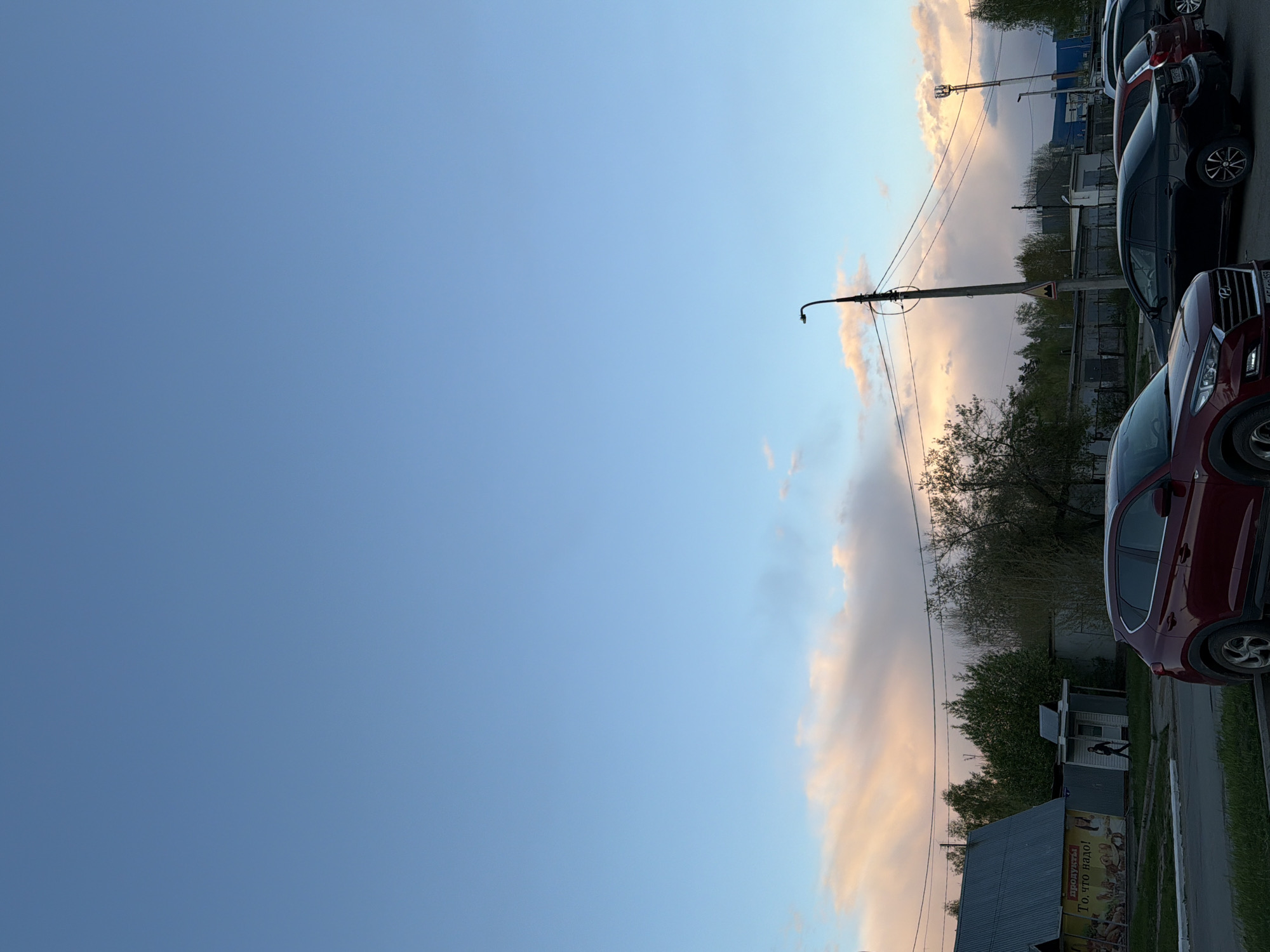 A sideways view shows the blue sky over the city of Chaikovsky, Russia. On the right side of the image, a row of parked cars is visible. These cars are partially blocking the view of buildings and trees behind them. There is a lamppost extending into the sky with power lines strung across. Clouds are seen in the background.