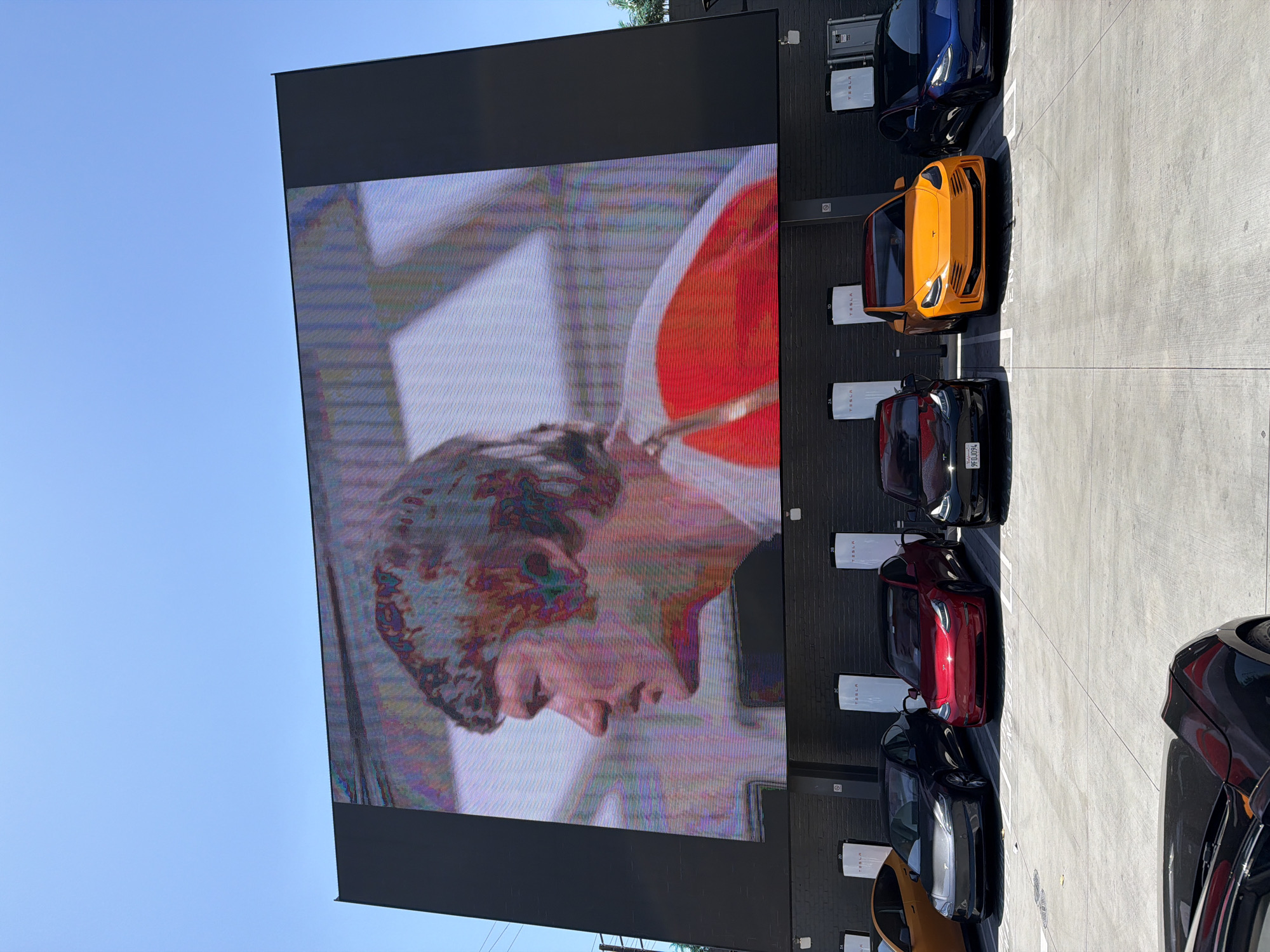 The image shows a large LED screen displaying a close-up of a person's face. The screen appears to be located outdoors, likely near a Tesla charging station, given the presence of Tesla vehicles and charging units parked along the wall to the right of the screen. The sky is visible above the screen and appears to be clear and blue. The location is in West Hollywood, United States of America.