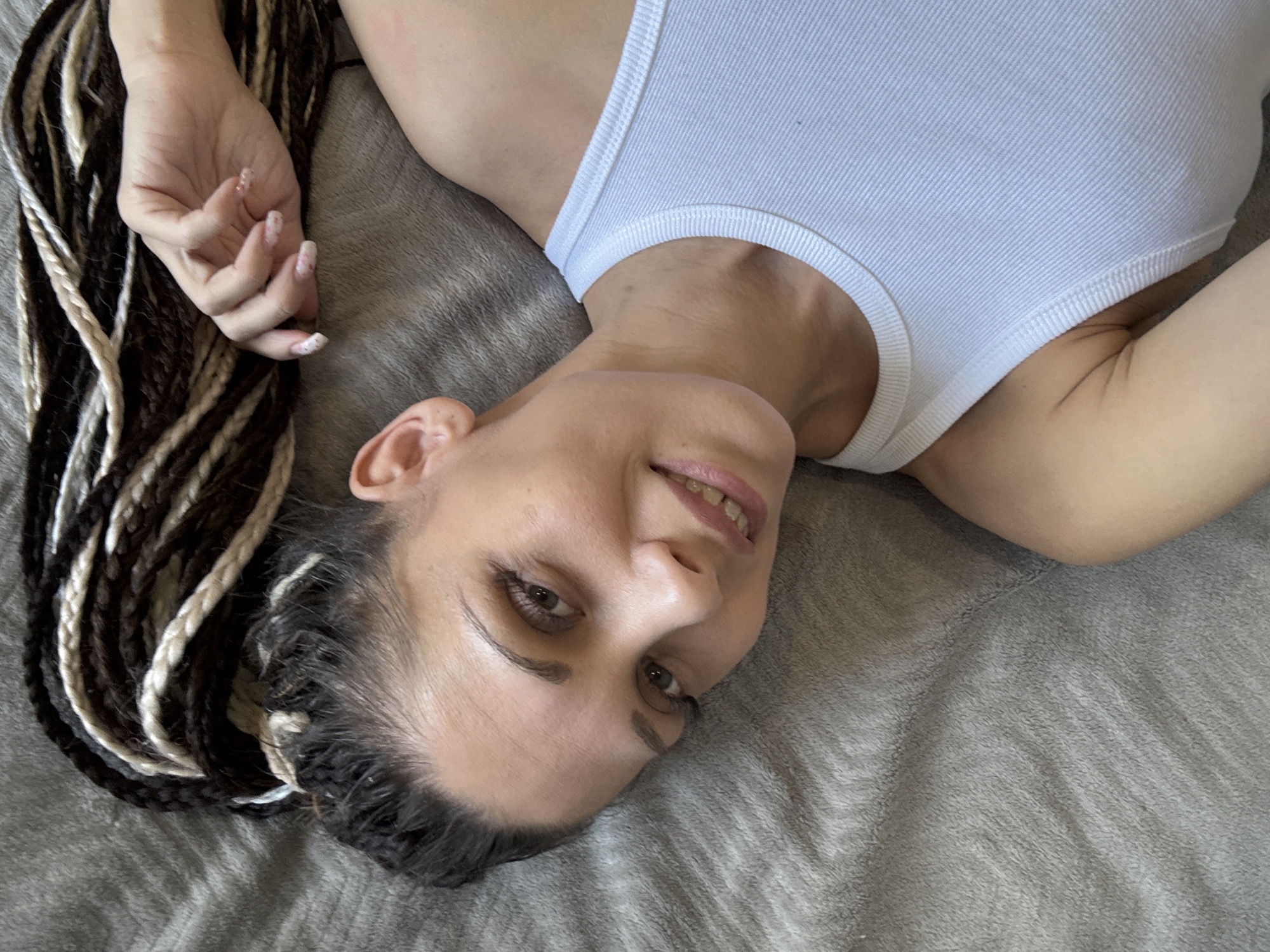 A woman with brown and blonde braided hair is lying on a gray blanket. She is wearing a white tank top and her right arm is visible. The location is Unknown, Unknown.
