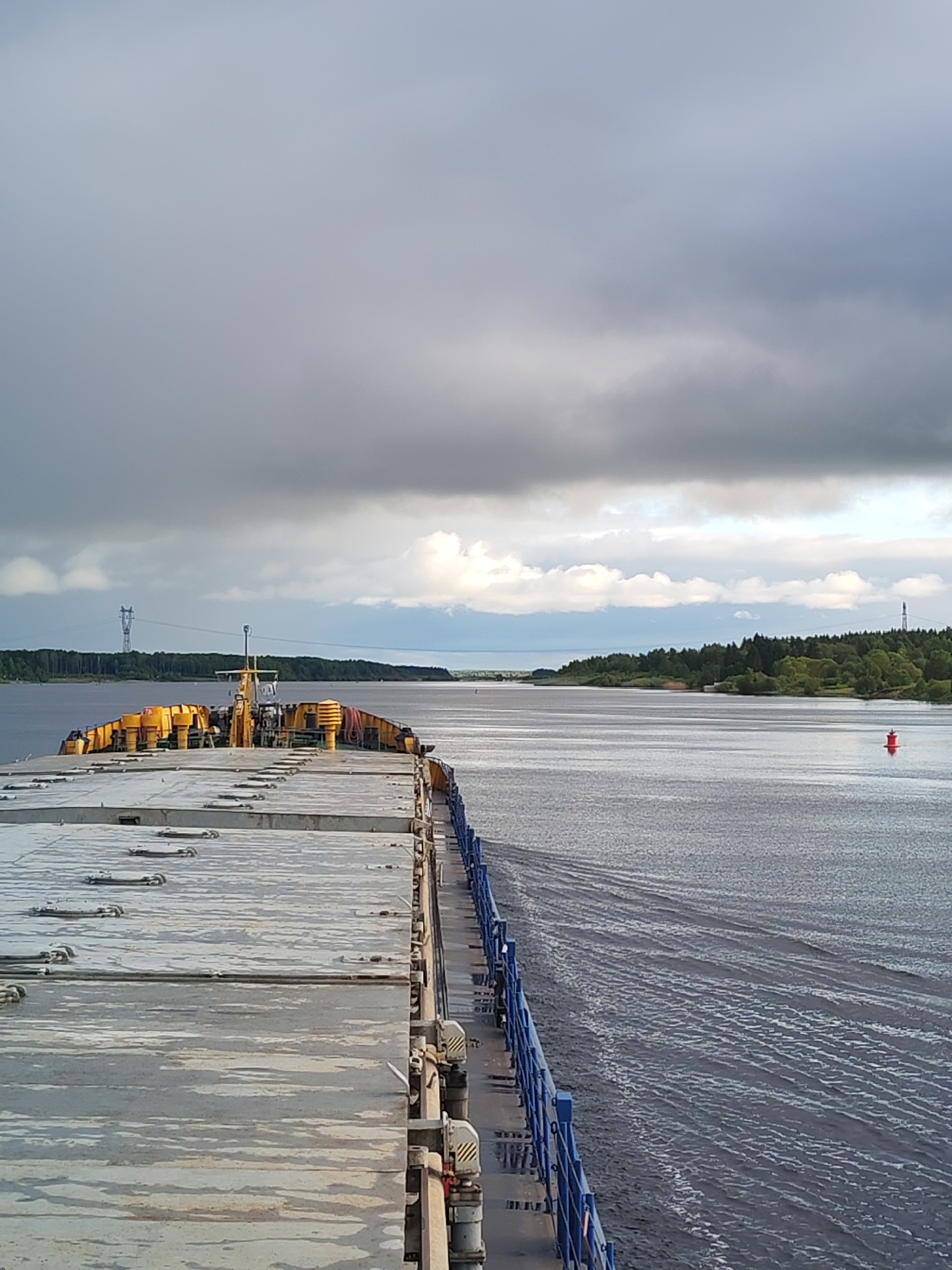 The image shows a view from a ship moving on a body of water, likely a river or wide canal, with land visible on both sides. The ship has a large, flat deck with gray, weathered surfaces and yellow equipment towards the bow. A blue railing runs along the starboard side of the ship.
The water is dark with ripples and wakes indicating movement. In the distance, a red buoy marks the channel. The land is covered in green trees. A power line tower is visible on the left side of the image.
The sky is overcast with gray clouds. The image likely depicts a view from near the city of Нифантово, Russia.