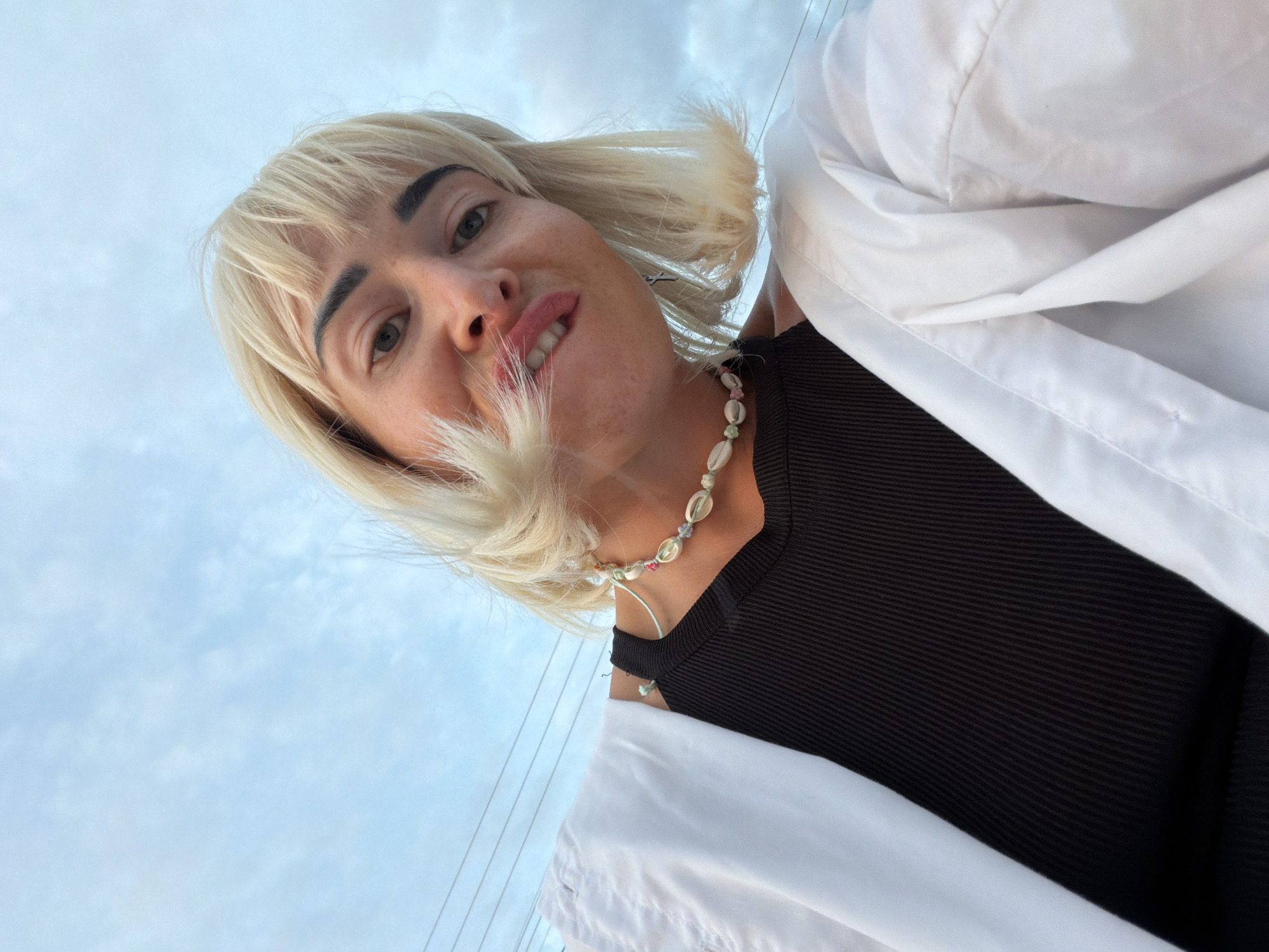 A woman with blonde hair and a white collared shirt over a black top looks at the camera. She is wearing a necklace made of shells and beads. The background is a cloudy sky with power lines visible. The image was taken outdoors, potentially in Sochi, Russia.
