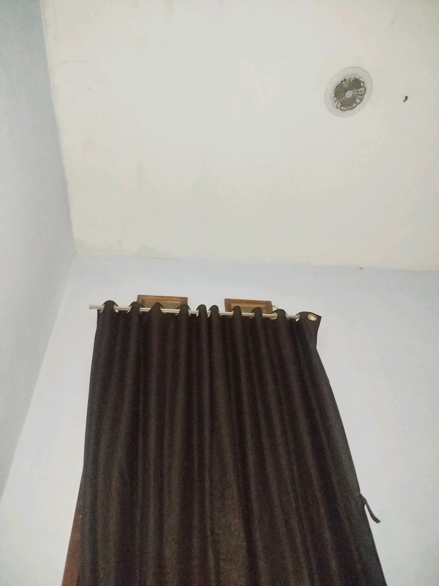 This is a low-angle, indoor shot of a dark brown curtain hanging from a white rod against a white wall and ceiling. A circular ventilation grate is visible on the ceiling, and what appears to be a fly or small insect is near it. The curtain is partially drawn, obscuring a window. The scene is located in a room in Babadan, Indonesia.