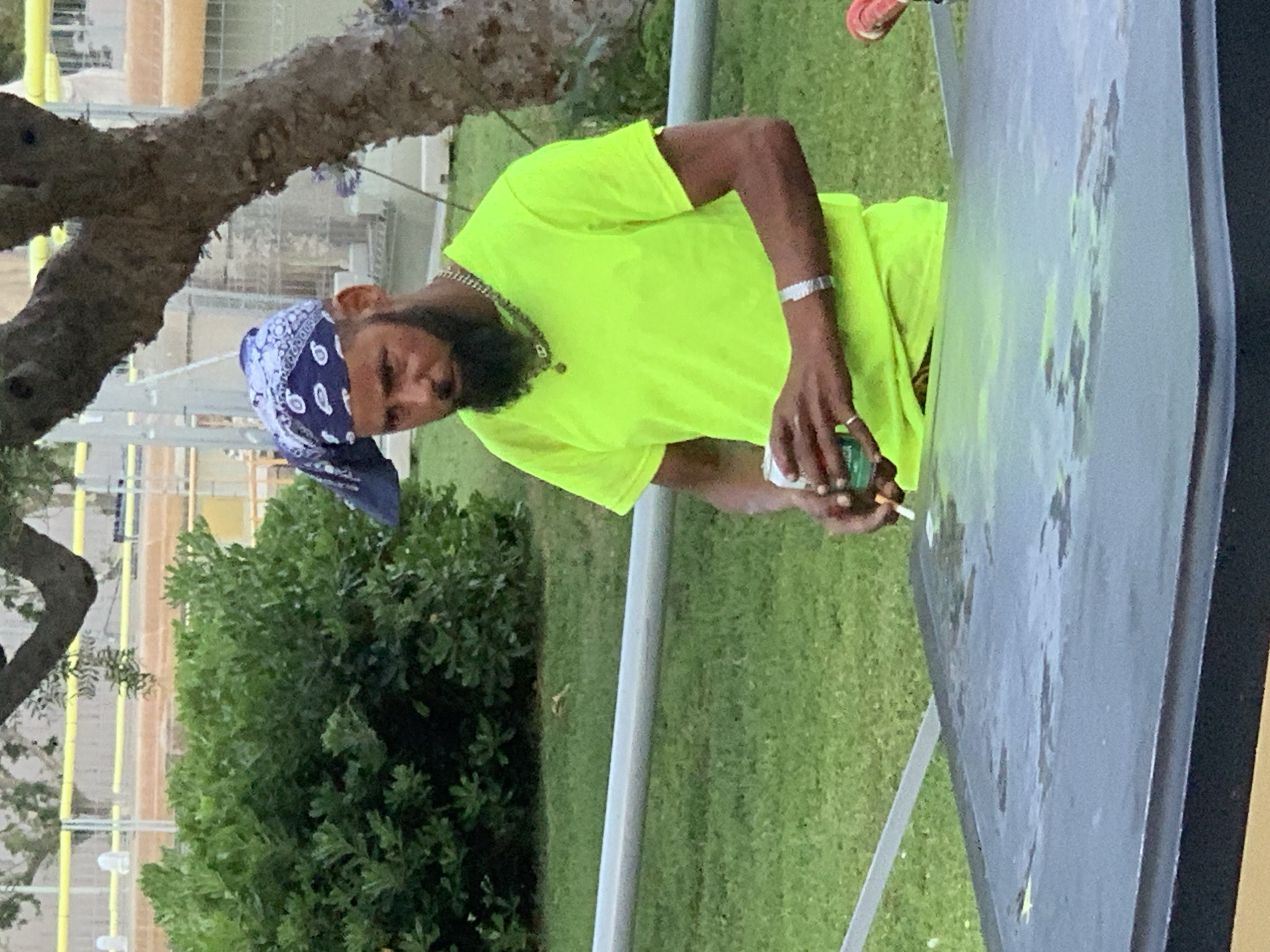 A man with a beard and a blue bandana on his head is standing outdoors. He is wearing a neon yellow t-shirt and a silver bracelet. He is holding a can in his left hand and a cigarette in his right hand. The man is standing in front of a dark-colored surface, possibly a table. In the background, there is green grass, a tree, and some fencing. There are no discernible landmarks or contextual clues to suggest the image was taken in Barcelona, Spain.