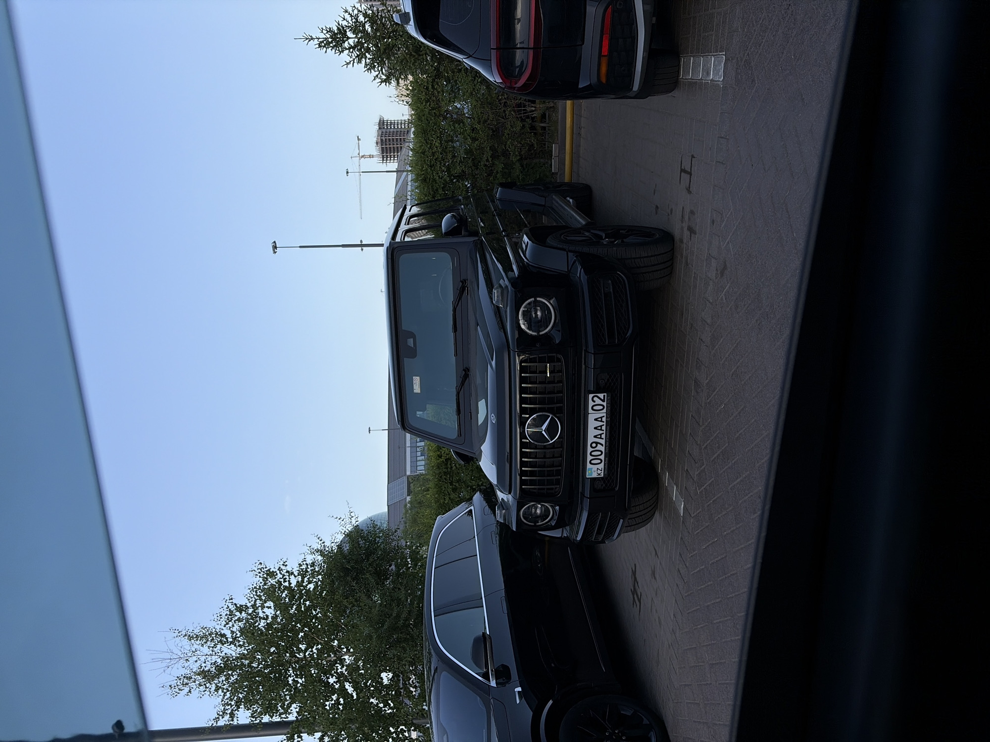 The image, captured outdoors during daytime, features a Mercedes-Benz G-Class (G-Wagen) parked on a paved surface, viewed from a slightly elevated angle. The dark-colored G-Wagen is positioned in the foreground, showcasing its prominent front grille with the Mercedes-Benz emblem and circular headlights.
The front license plate of the G-Wagen displays "KZ" in blue, followed by "009AAA 02". Smaller text, "Qazaqstan" and "www.gov.kz", is also visible. "KZ" indicates Kazakhstan; while "02" typically corresponds to the Aktobe region, the provided context places the location in Astana, Kazakhstan.
Behind the G-Wagen, another dark SUV is partially visible, showing its rear section and taillights. A third dark vehicle is partially seen on the far left edge of the frame. The ground is covered with dark, brick-patterned paving.
In the mid-ground, green bushes and trees line the parking area. The background features a clear, light blue sky, against which multiple construction cranes and the framework of a building under construction are visible. A street light pole stands in the background. The overall scene suggests a clear, pleasant day in an urban or developing area, with no human activity visible apart from the parked vehicles.