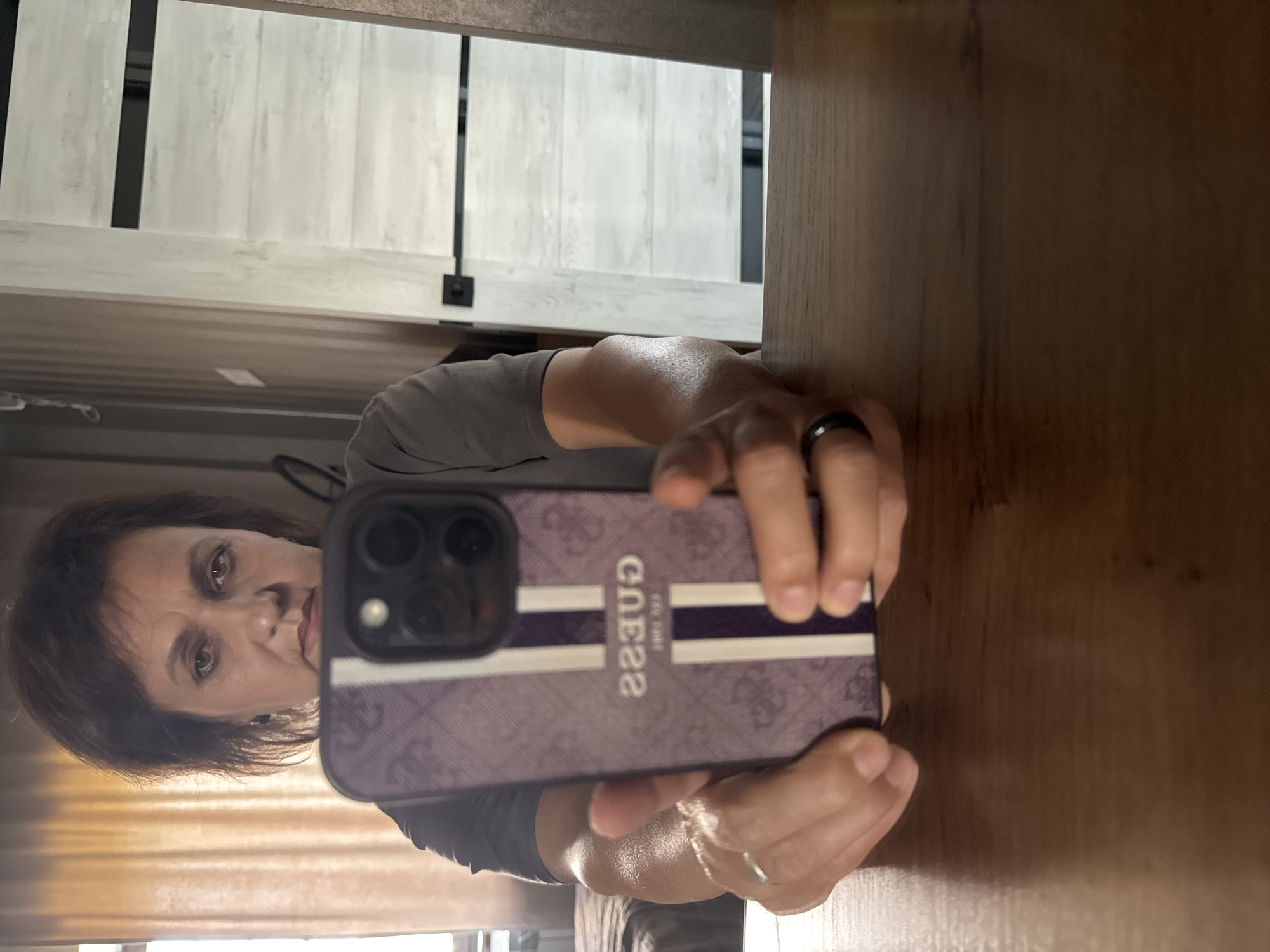 In this waist-up, indoor, mirror selfie, a woman with dark, layered hair and brown eyes is holding a purple phone with a Guess logo. She is wearing a grey t-shirt and a ring on her left ring finger and a black ring on her right ring finger. The background shows a cream-colored door with black horizontal accents, a sheer curtain, and a bed. The scene appears to be a bedroom or dressing area.