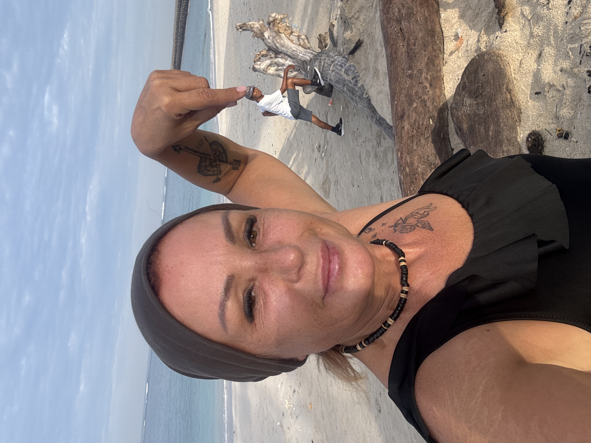 A selfie of a woman on a beach. The woman is fair-skinned with light blonde hair, wearing a gray head covering, a black top, and a black beaded necklace. She has tattoos on her left arm and right collarbone area. She is smiling at the camera and holding her left hand up, appearing to "pinch" a man in the background with her fingers.
In the background, a man is standing next to a large piece of driftwood. He is dark-skinned, wearing a white shirt, gray shorts, and a gray hat. The location is a sandy beach with the ocean and sky visible in the background. The ocean appears calm. Based on the landscape and the presence of driftwood on the beach, the city of Denpasar, Indonesia, cannot be confirmed.
