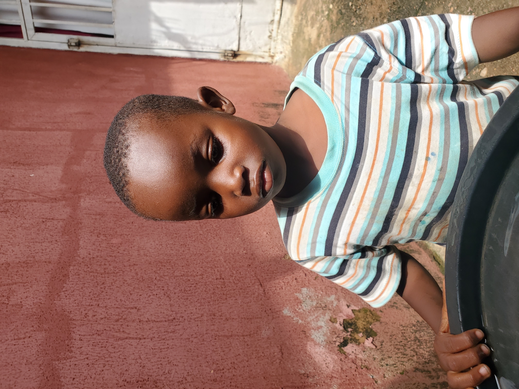 A young child with short hair, wearing a striped t-shirt with blue, orange, and gray stripes, is shown in a close-up portrait. The child is looking downwards with their eyes closed, their face slightly angled. The t-shirt has a light blue collar. The child's left hand is visible, resting on a dark, round object, possibly a container. The background features a reddish-brown textured wall and a white structure with horizontal slats, suggesting an outdoor setting, possibly a home or building exterior. Sunlight casts shadows, indicating it is daytime. The scene appears to be set in Jos, Nigeria. There is no visible text in the image.