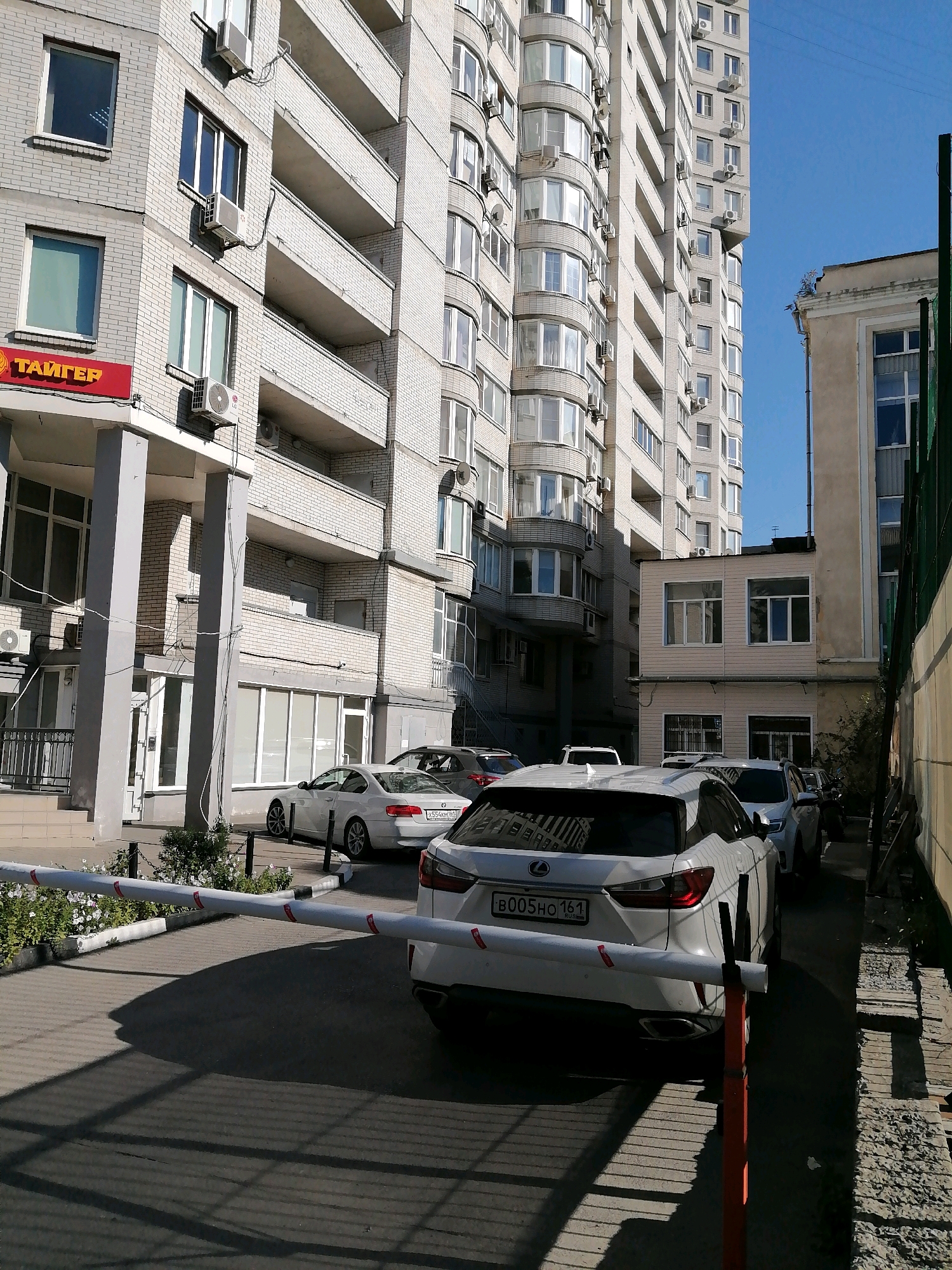 The image depicts an outdoor scene in Rostov-on-Don, Russia, featuring a modern apartment building with a light-colored brick facade and multiple balconies. In the foreground, a white Lexus SUV is parked behind a white barrier with red markings. The SUV has a license plate that reads "B005HO 161 RUS." Several other vehicles are also visible, including a white sedan and a dark-colored SUV. A portion of a beige building with windows is visible to the right of the apartment building, and a green fence runs along the right edge of the image. The ground is paved with cobblestones. The sky is clear and blue. The scene appears to be a parking area or courtyard adjacent to residential buildings.