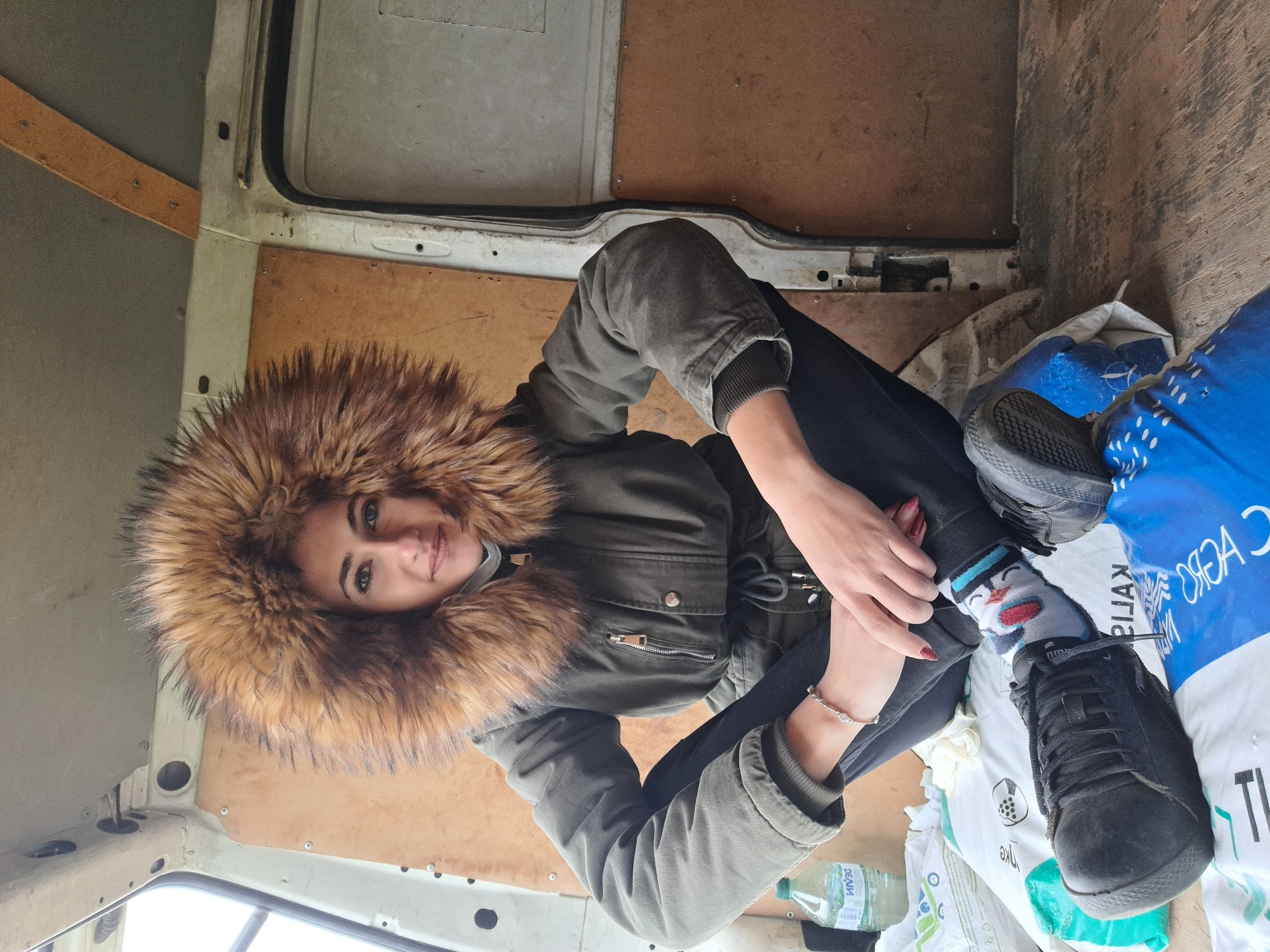 A woman is sitting inside a van in Negovan, Bulgaria. She is wearing a green winter jacket with a fur-lined hood, black pants, and black shoes with a snowman sock on one foot. She is sitting on a blue and white bag labeled "Kalia Agro" and other white bags. A bottle of water is near her. The interior of the van is unadorned, with plywood walls and a utilitarian appearance. The lighting suggests it is daytime.